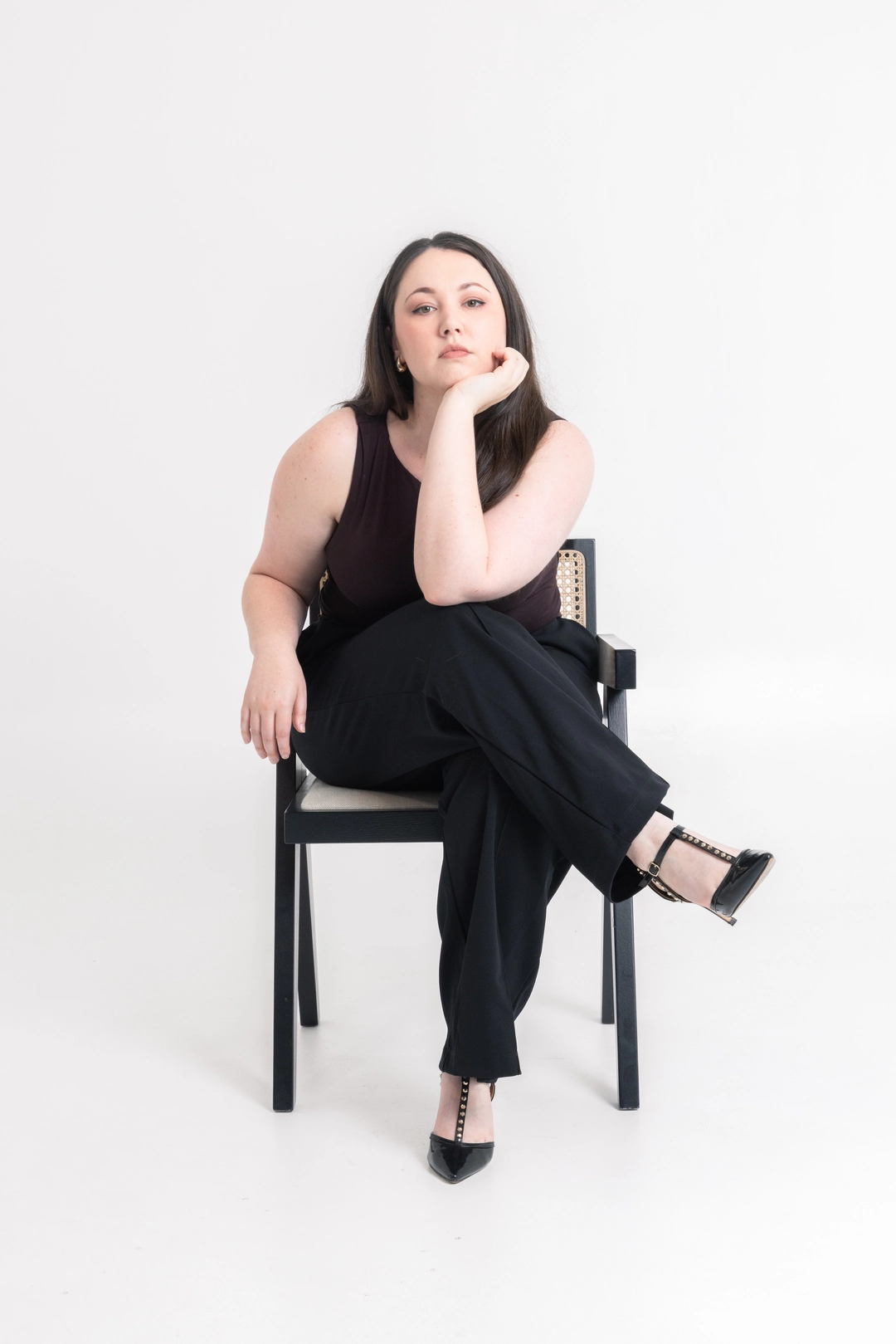 Woman with long dark hair wearing a sleeveless black top and black pants standing against a plain light background.