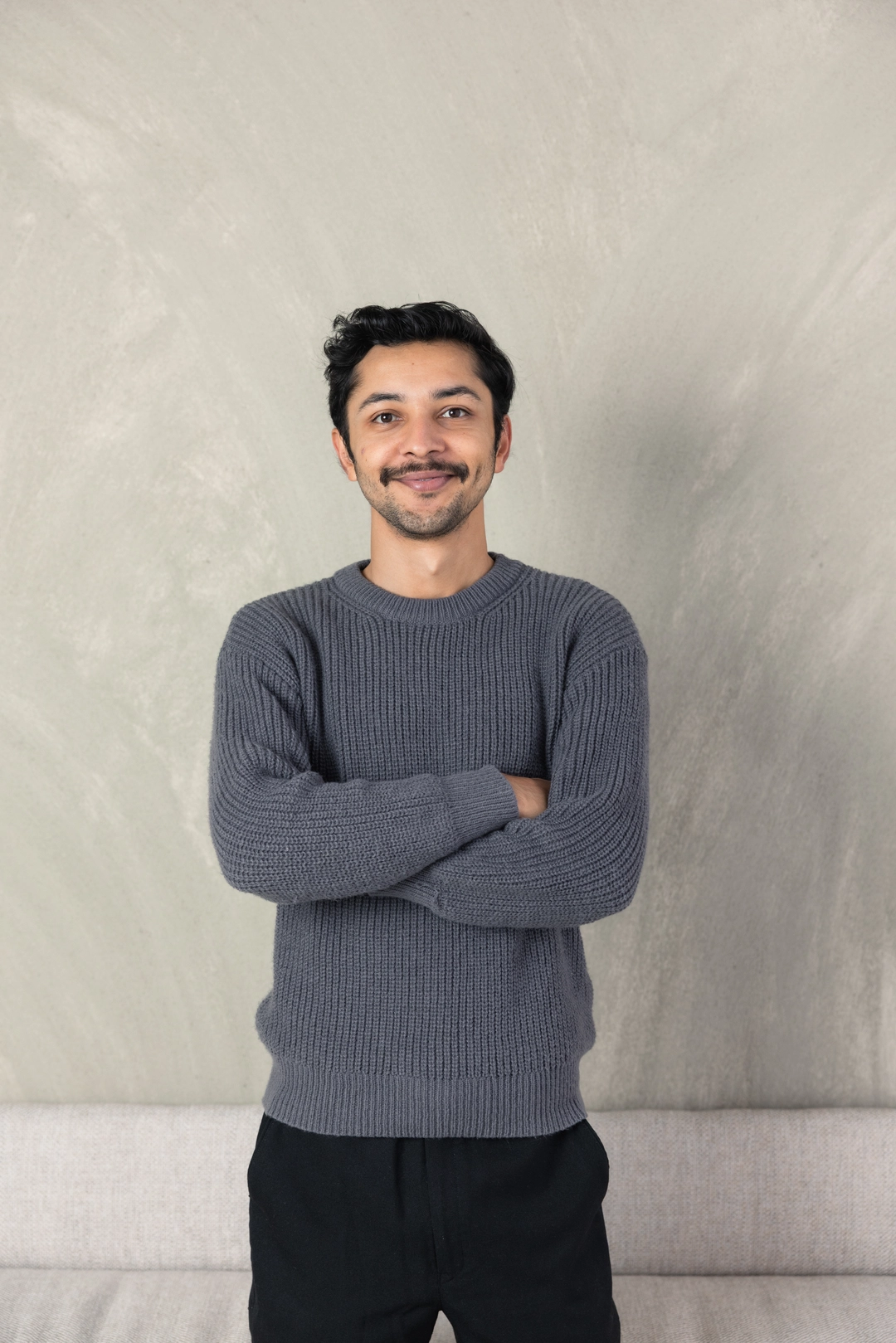 Aman Hirani, website director at Adcraft Studio, standing with his arms crossed and smiling against a textured grey wall.