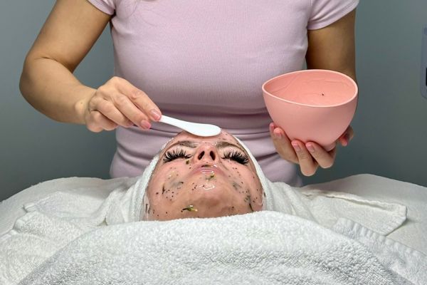 Person receiving a facial treatment with a facial mask applied, while a beautician in a pink shirt holds a bowl and applicator.