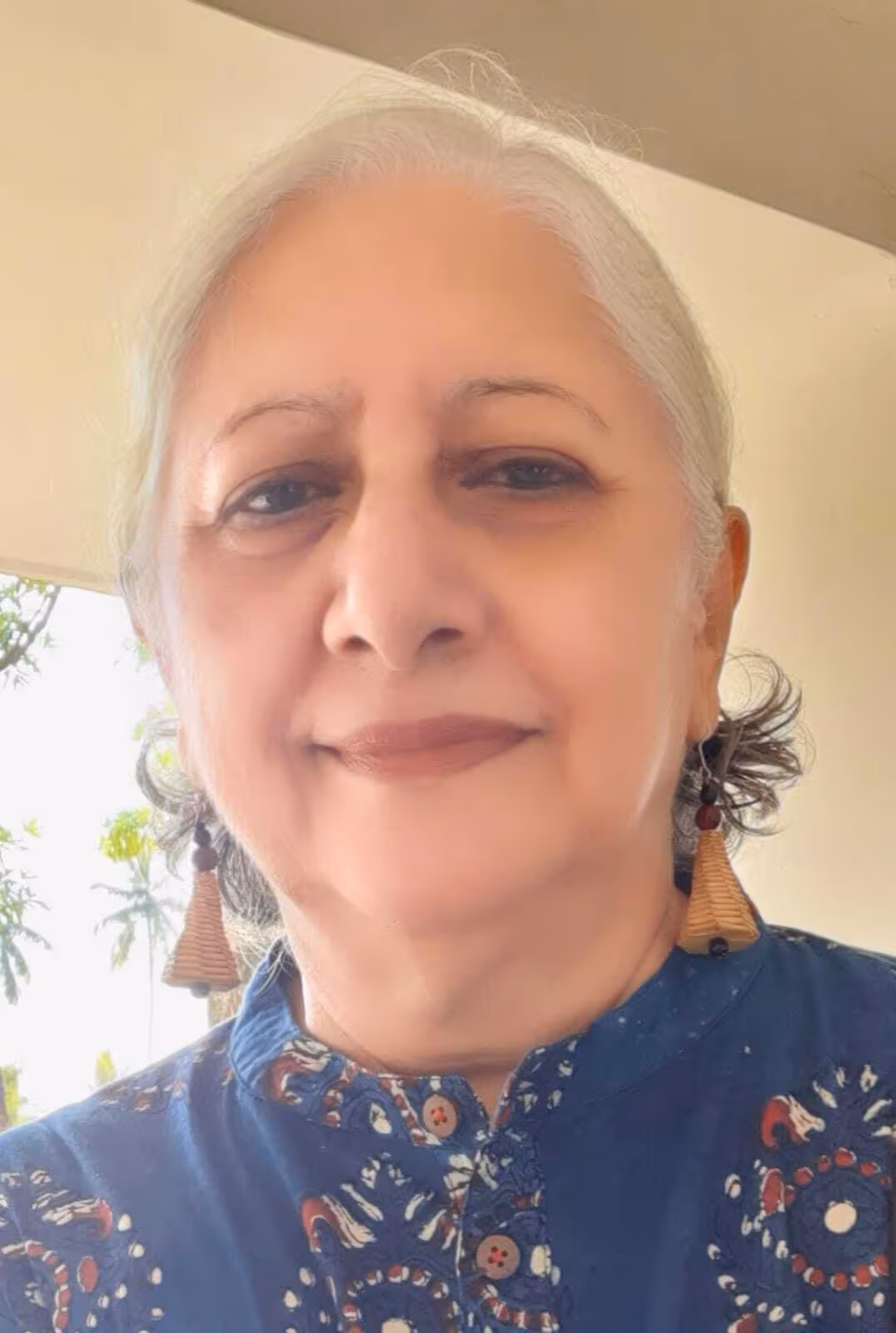 Smiling elderly woman with gray hair tied back, wearing patterned blue top and wooden earrings.