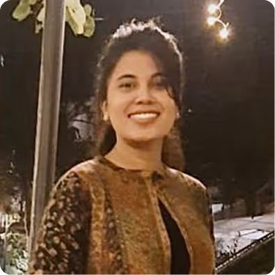 Smiling woman with dark hair wearing a patterned jacket, standing outdoors at night with lights in the background.