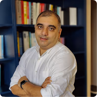 Man with short hair wearing a white shirt and smartwatch, standing with arms crossed in front of a dark bookshelf filled with books.
