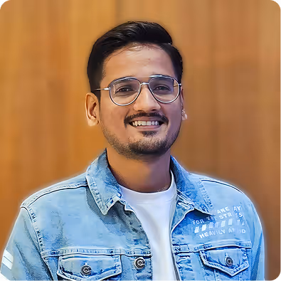 Smiling man with short black hair and glasses wearing a light blue denim jacket over a white shirt, standing against a blurred brown background.