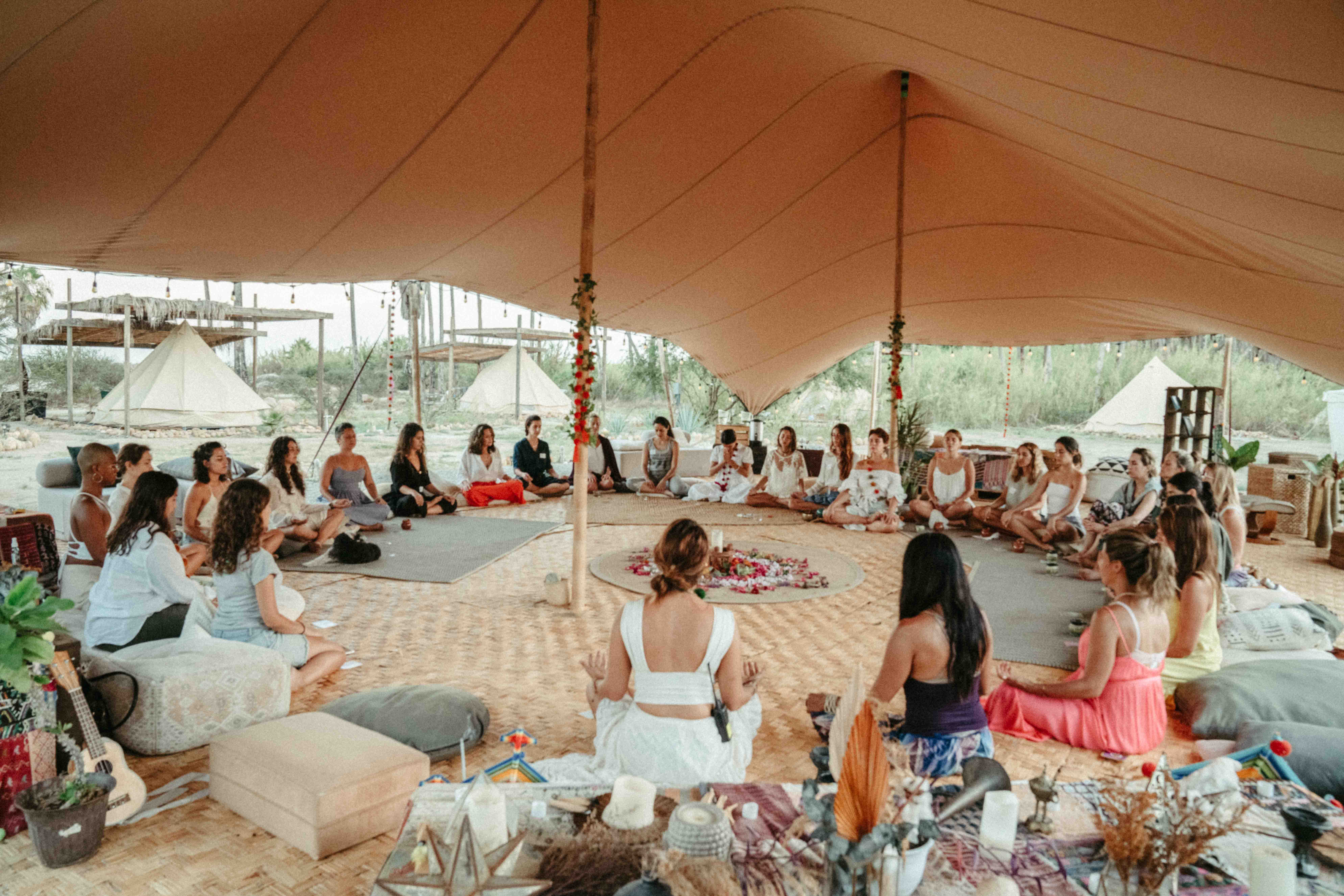 Group of people sitting in a circle inside a large tent, participating in a meditation or group session with a decorated central area.