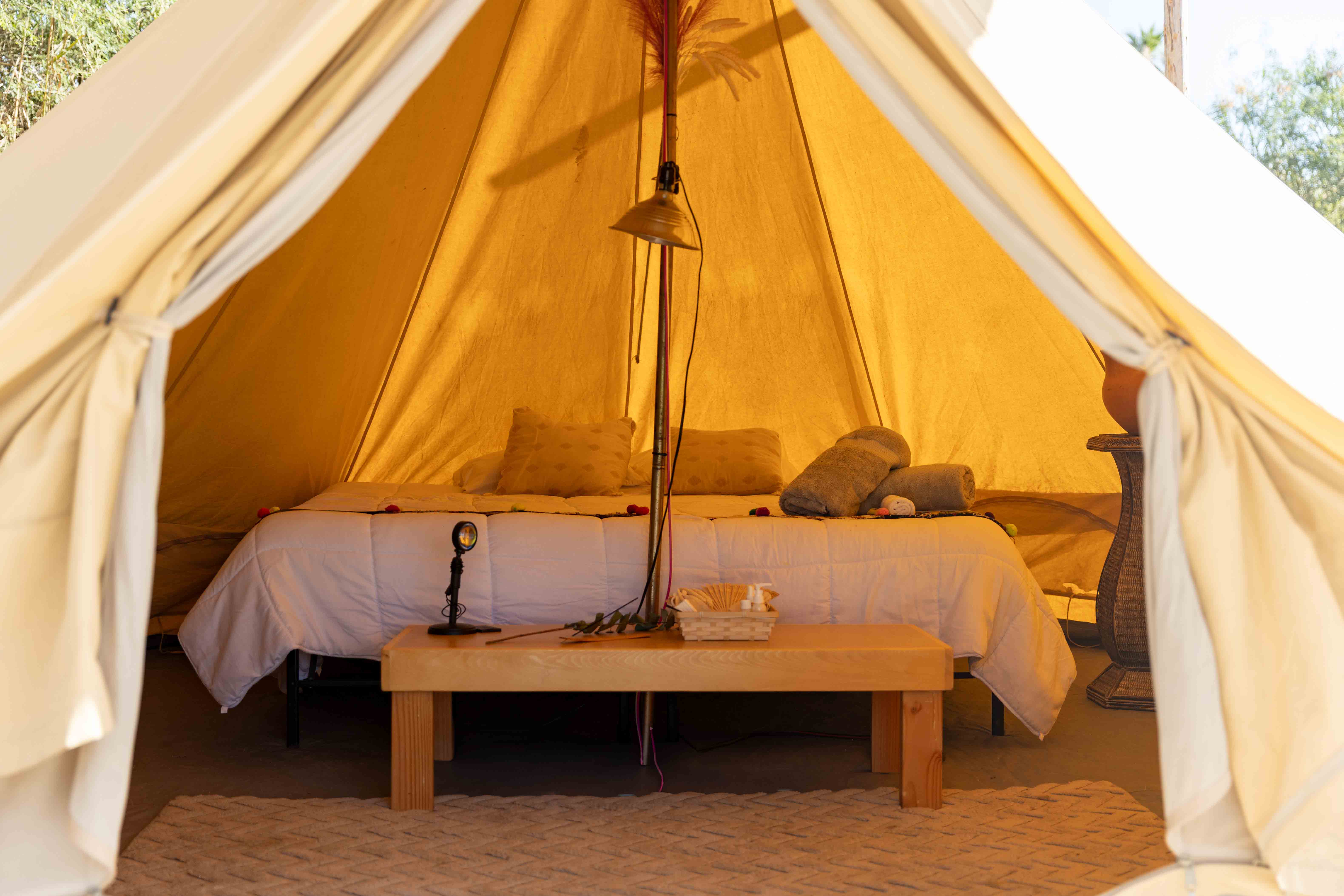 Inside a beige canvas tent with a made bed, rolled towels, pillows, and a wooden table with toiletries and a lamp.
