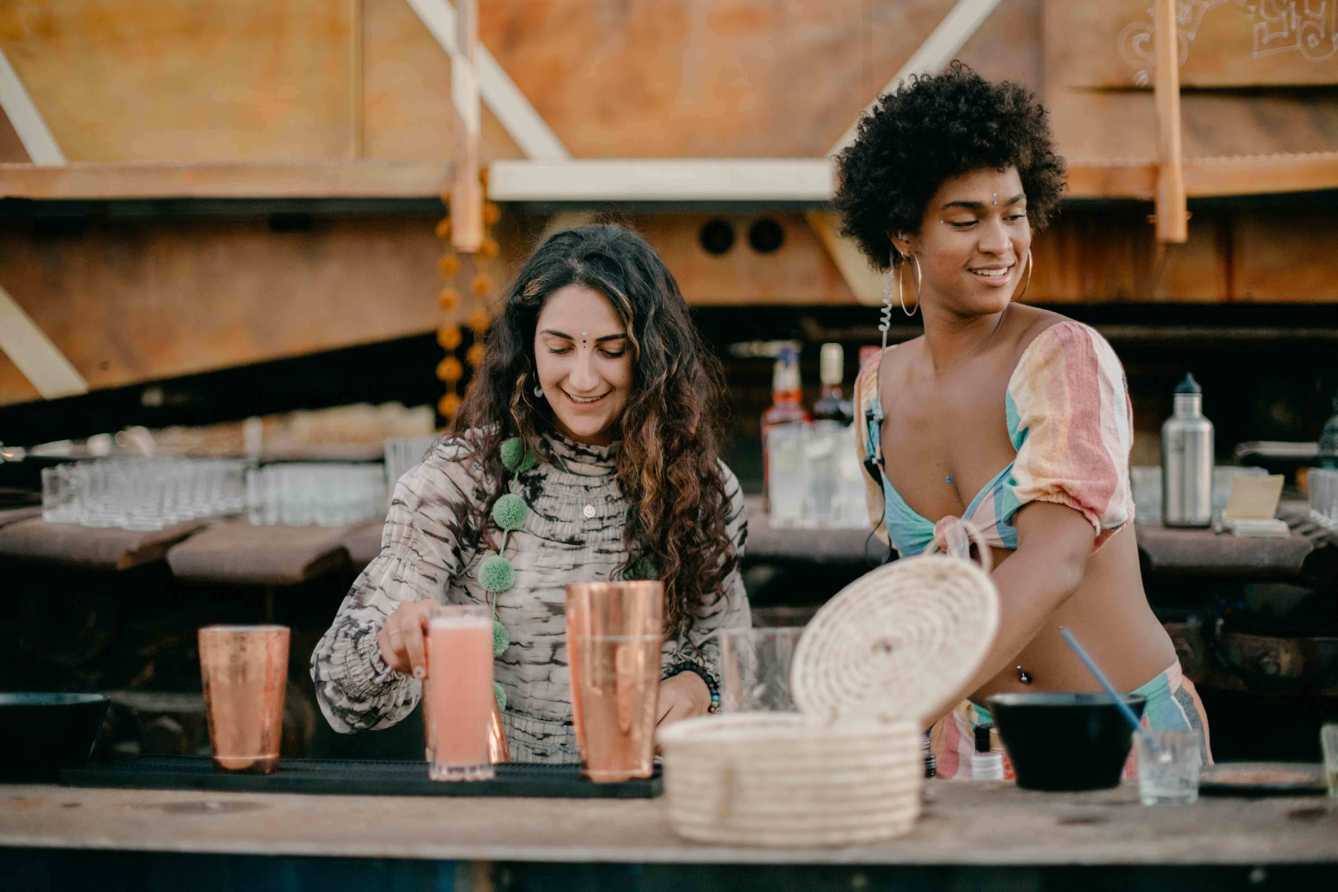Two women smiling behind a bar, one holding a pink drink and the other reaching for something on the counter.