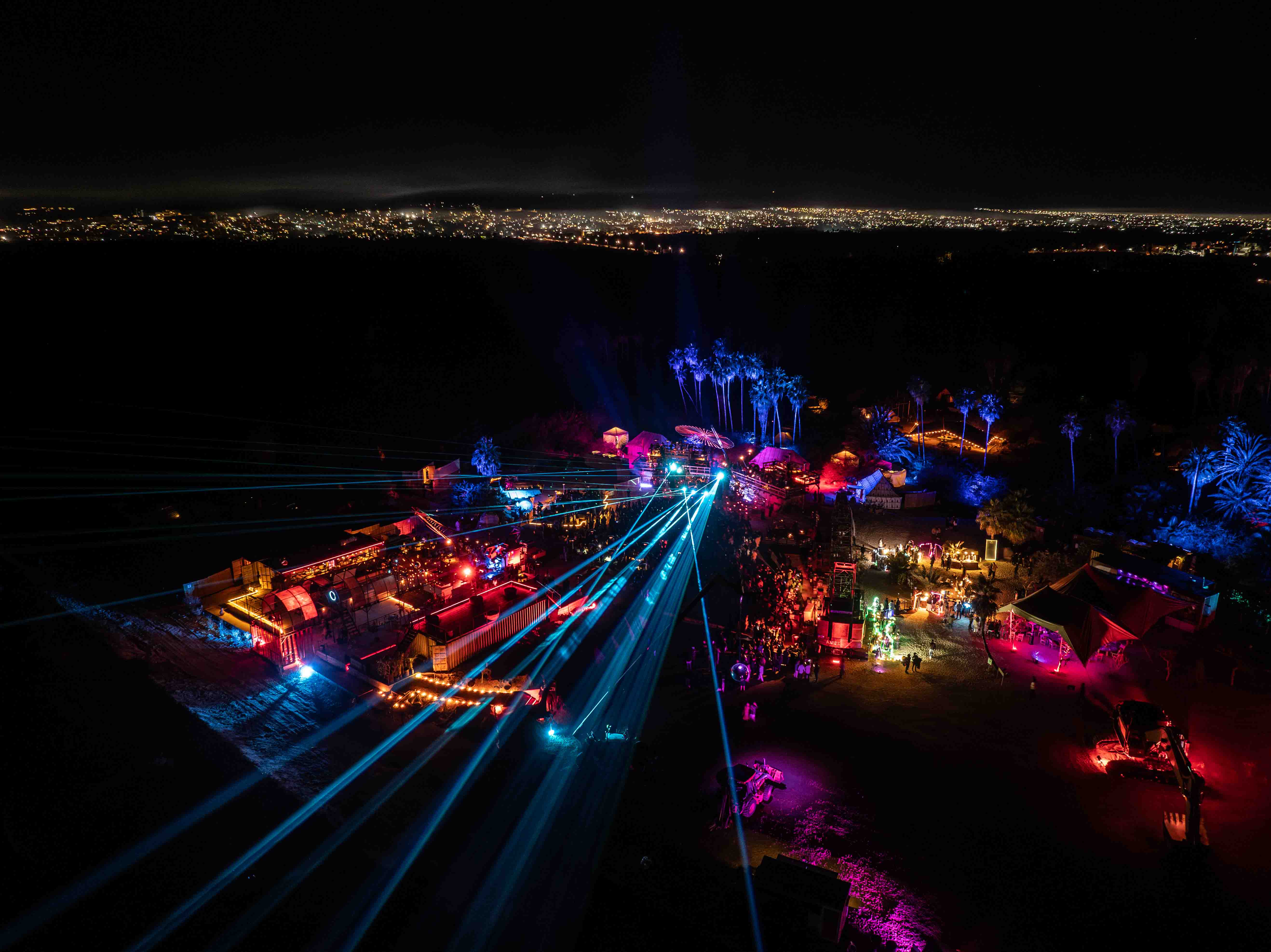 Aerial view of a nighttime outdoor event with colorful lights, laser beams, palm trees, and a crowd of people.