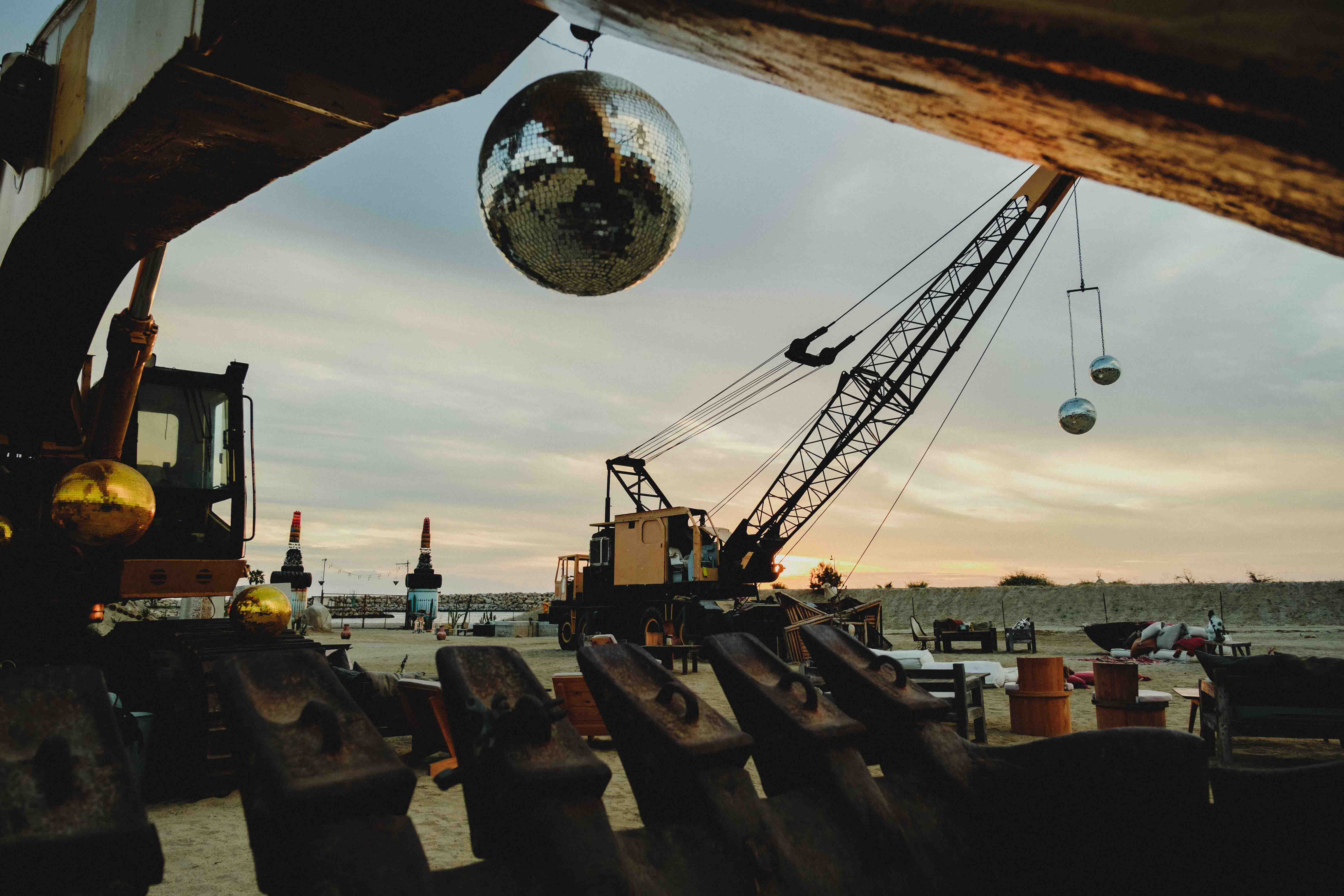 Large construction crane outdoors at sunset with disco balls hanging from it and scattered seating and furniture on sandy ground.