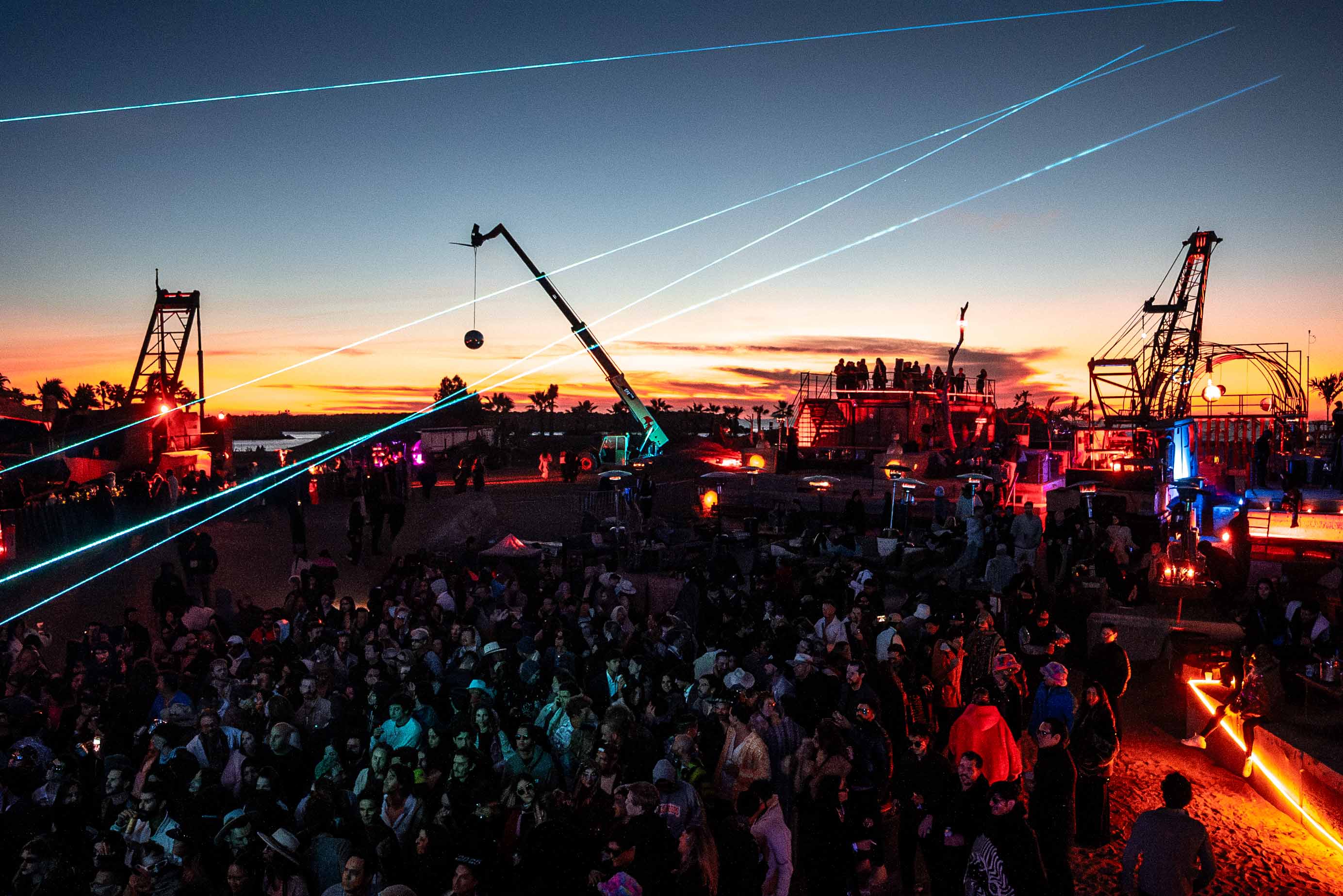 Large crowd gathered at an outdoor event at sunset with cranes, laser lights, and illuminated structures.