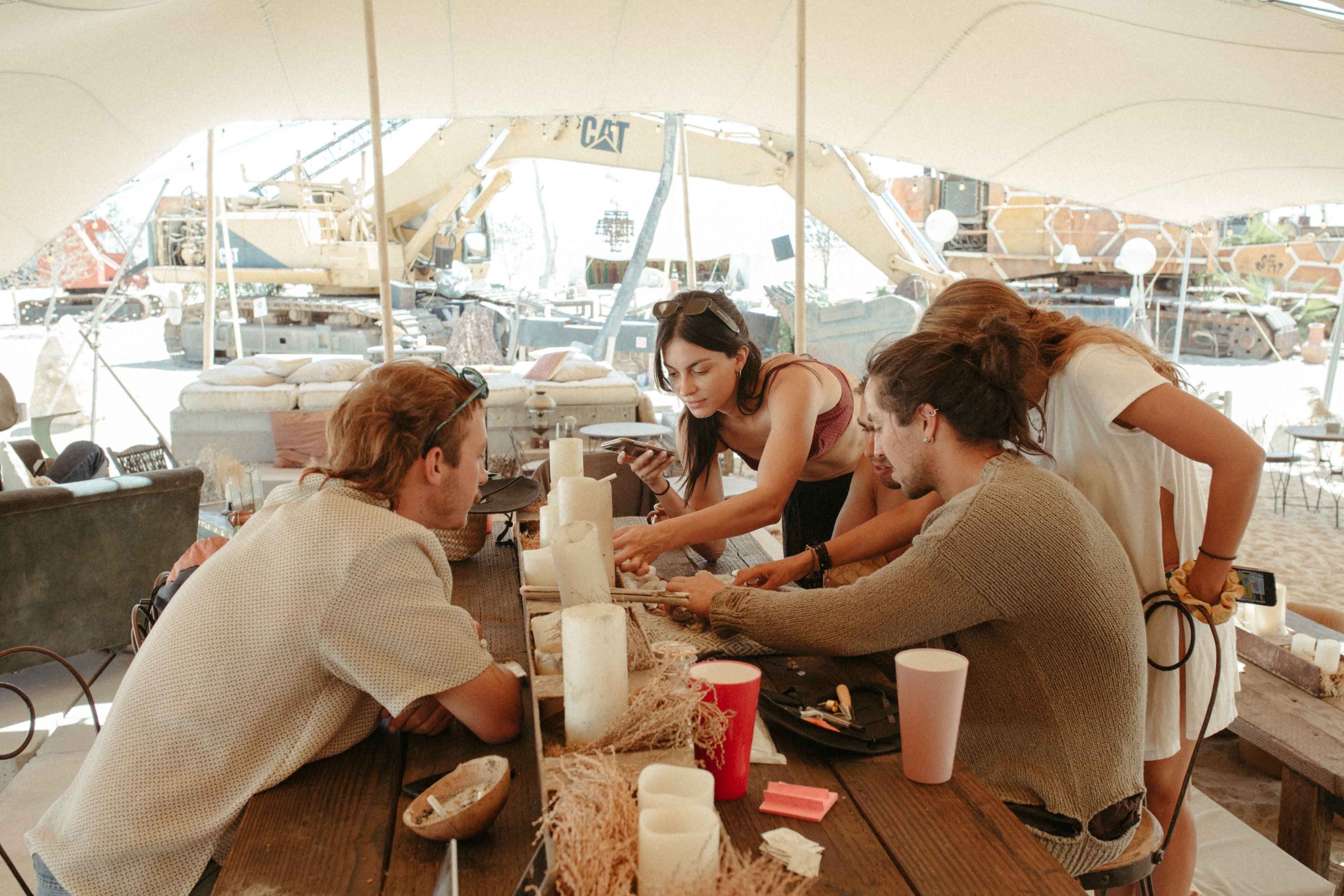 Four young adults sitting and standing around a wooden table under a canopy, engaged in a discussion with construction equipment visible in the background.