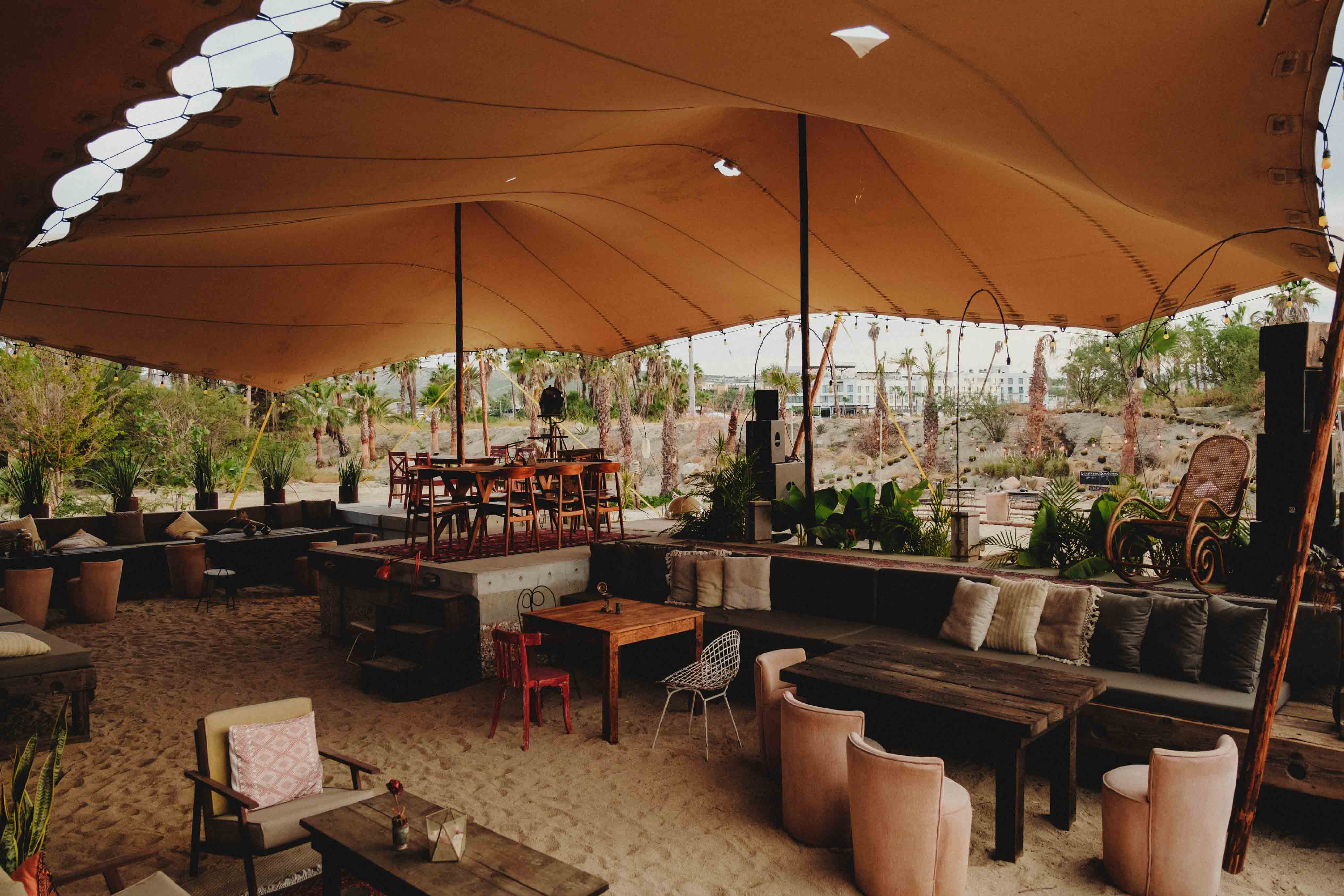 Outdoor lounge area with mixed wooden and upholstered seating under a large beige canopy on sandy ground, surrounded by palm trees.