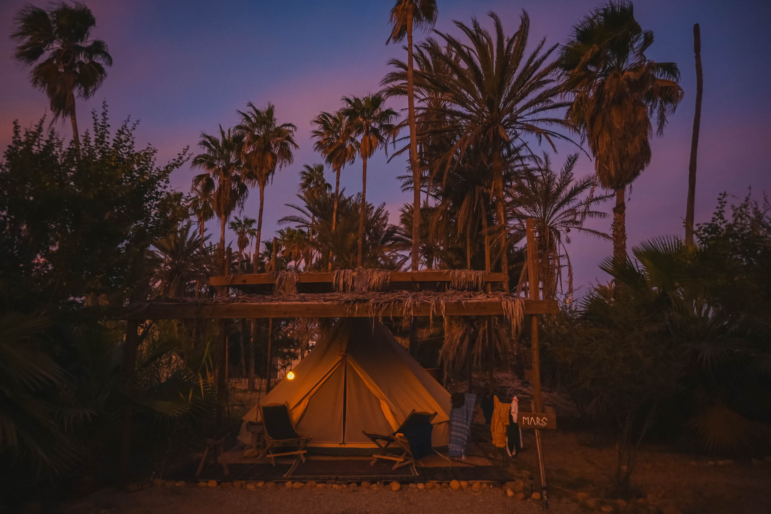 A cozy tent illuminated at dusk under palm trees with two chairs outside and a wooden sign labeled Mars.