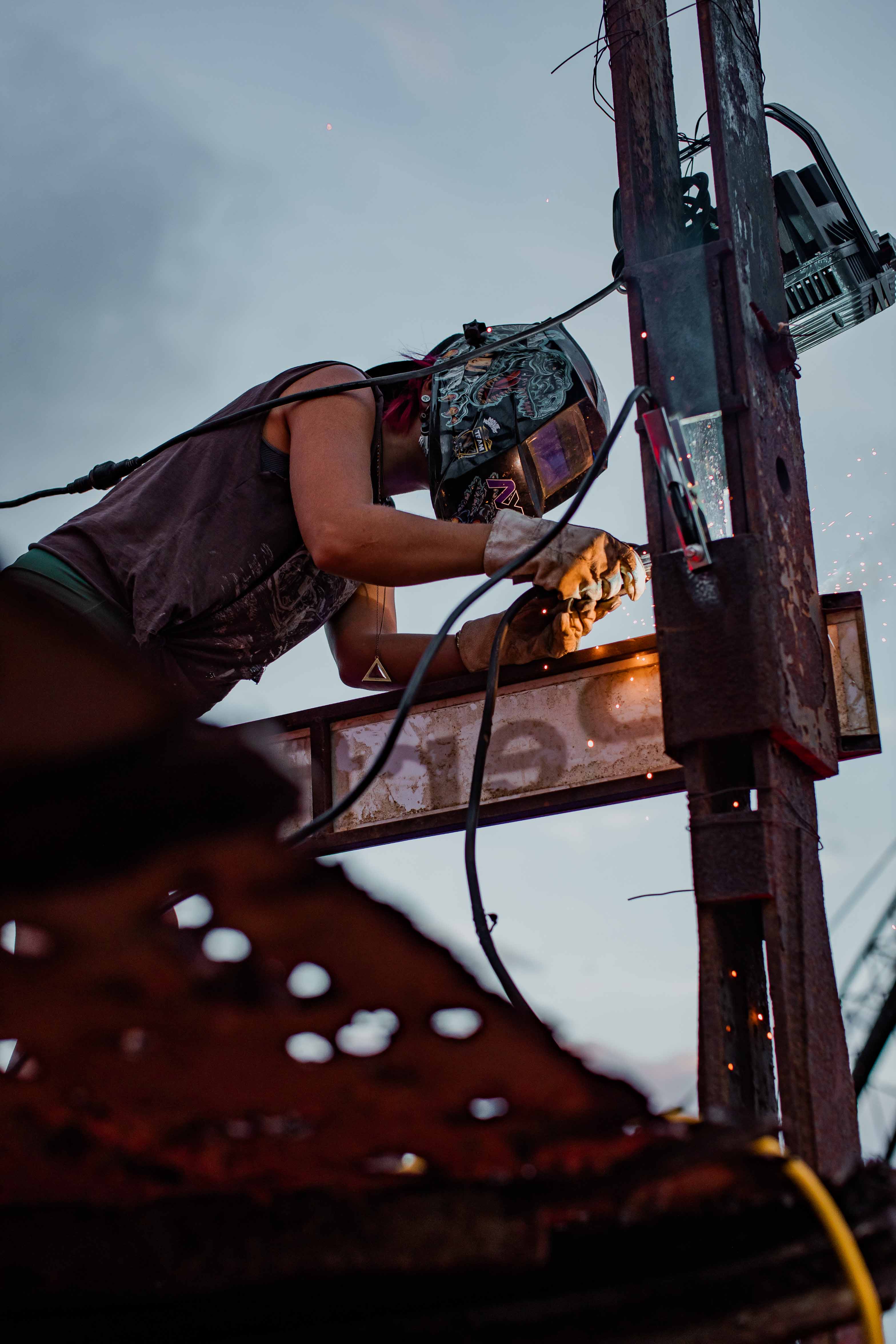 Person wearing a decorated welding helmet and gloves welding metal on a pole with sparks flying against a dusk sky.