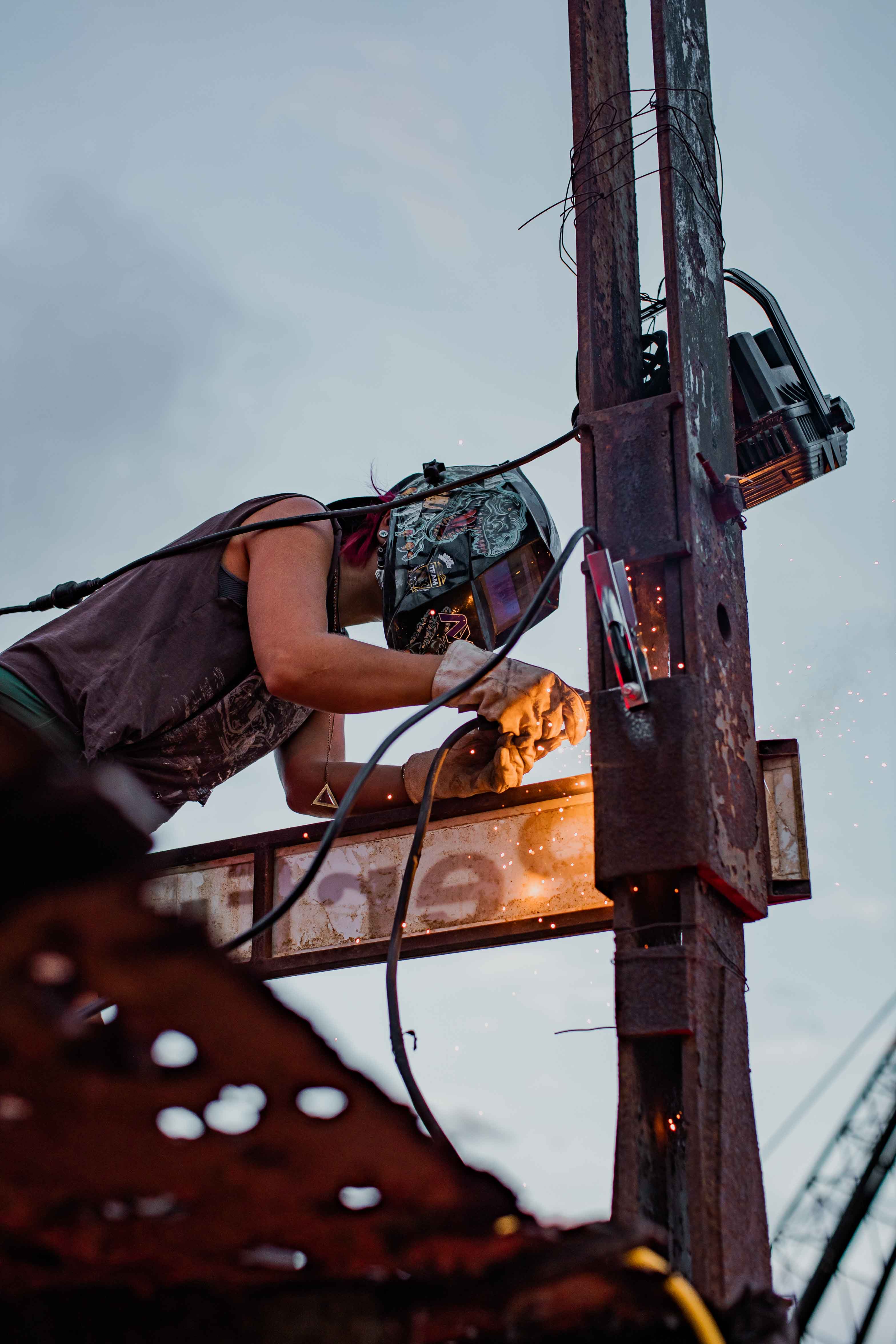 Person wearing protective welding helmet and gloves welding metal on a rusty pole with sparks flying.