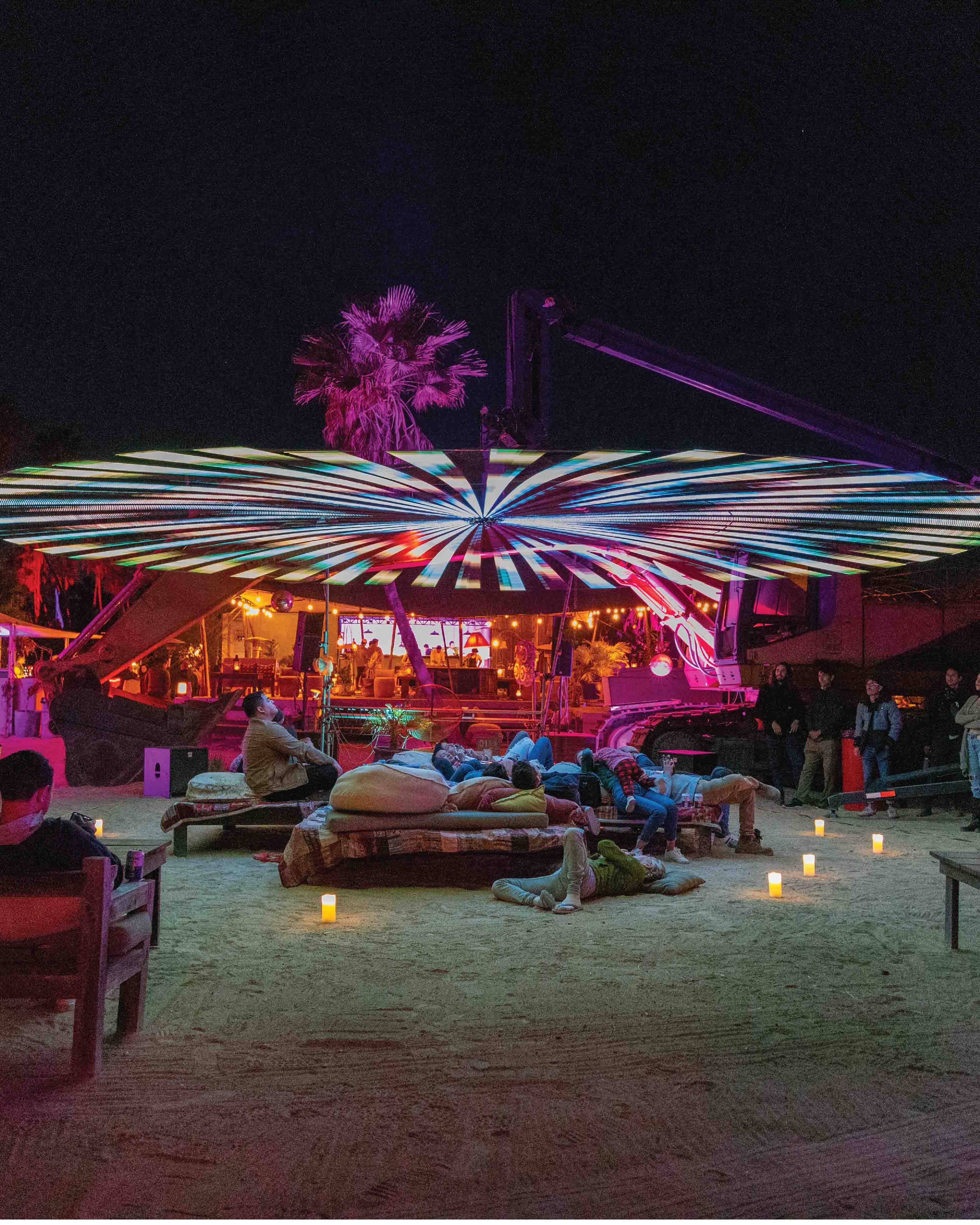 People relaxing on cushions and benches under a large circular LED-lit canopy at night, with colorful lights and candles on sandy ground.