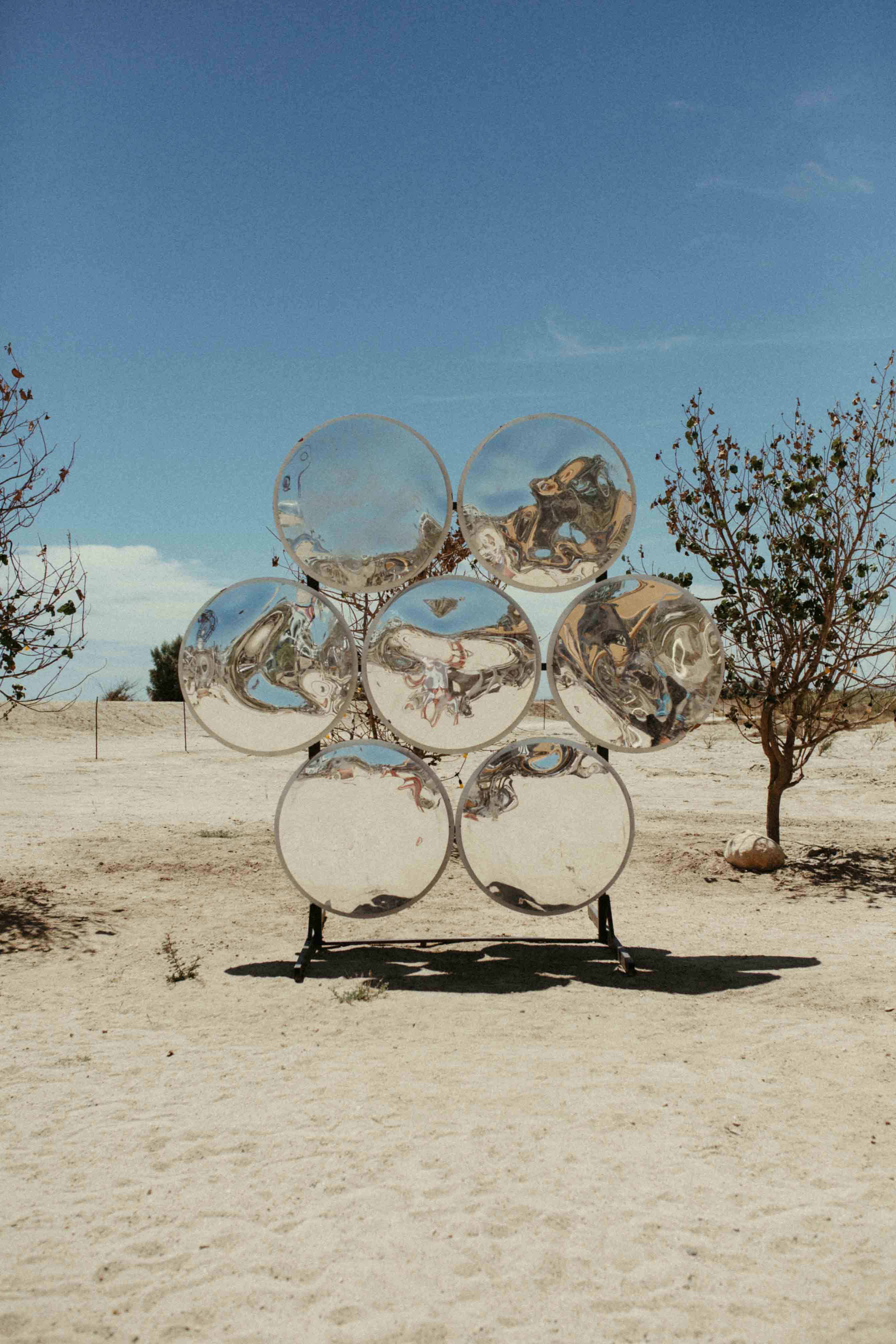 Seven round, reflective metal discs arranged in a flower shape on a sandy desert ground with two small trees nearby under a clear blue sky.