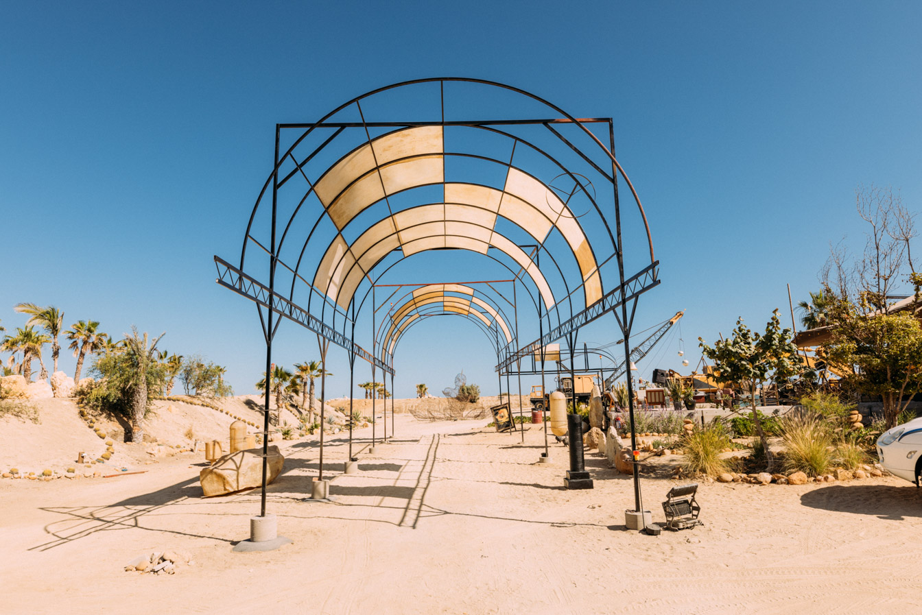 Metal archway structure casting shadows on a sandy path with palm trees and desert plants under a clear blue sky.