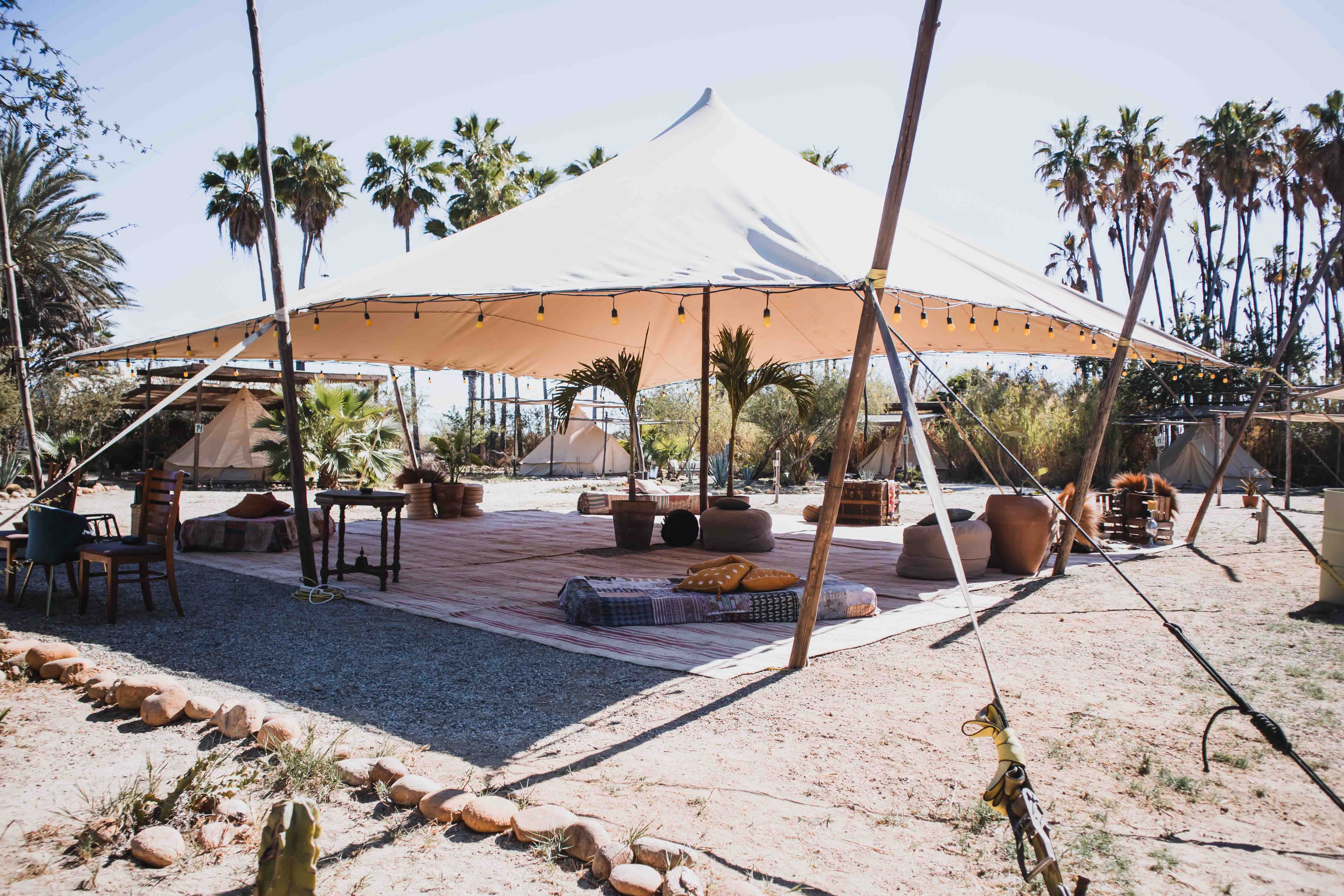 Open outdoor tent with seating cushions, rugs, potted plants, and string lights under a clear sky with palm trees in the background.