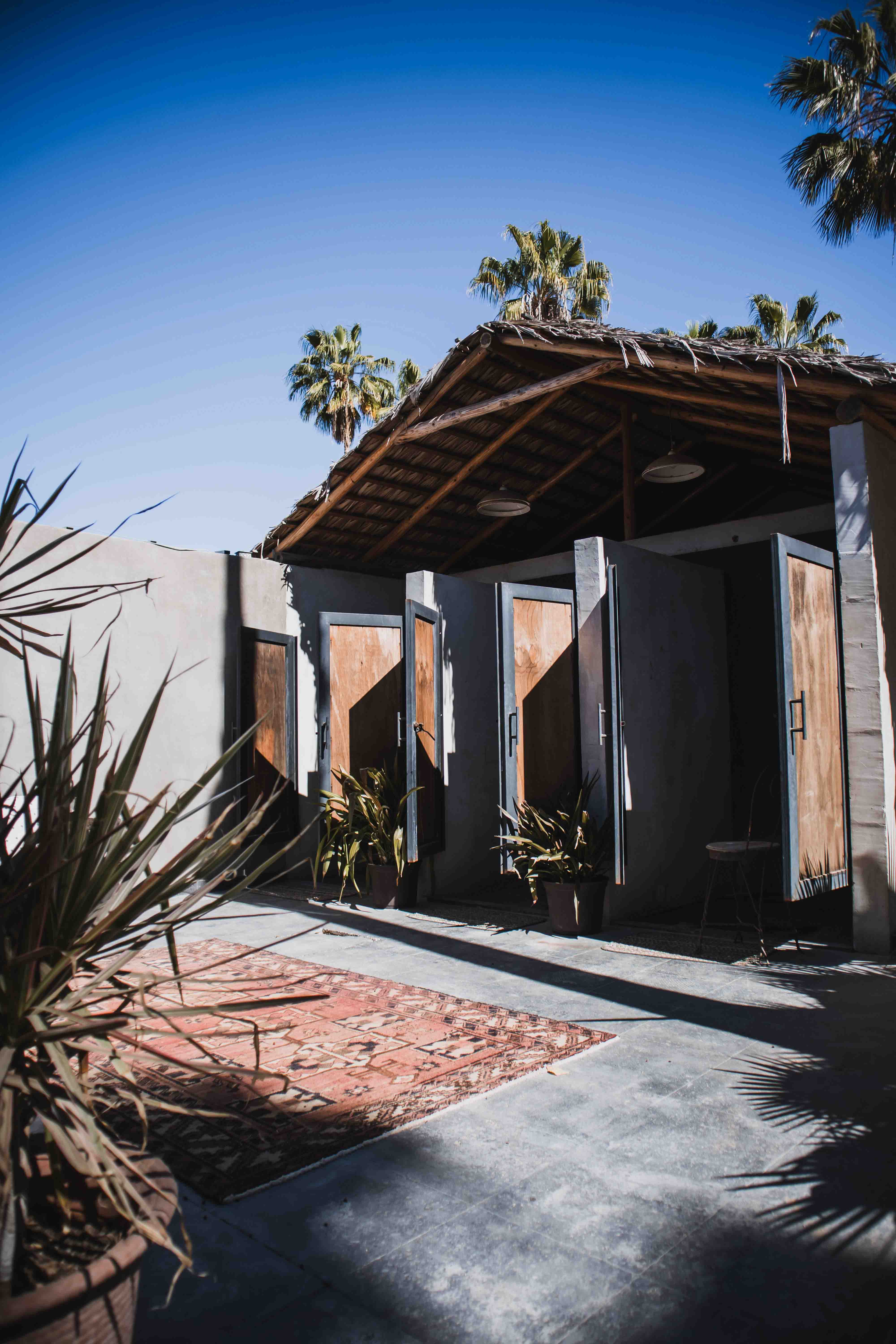 Outdoor area with wooden changing room doors, potted plants, a patterned rug, and a thatched roof under a clear blue sky.