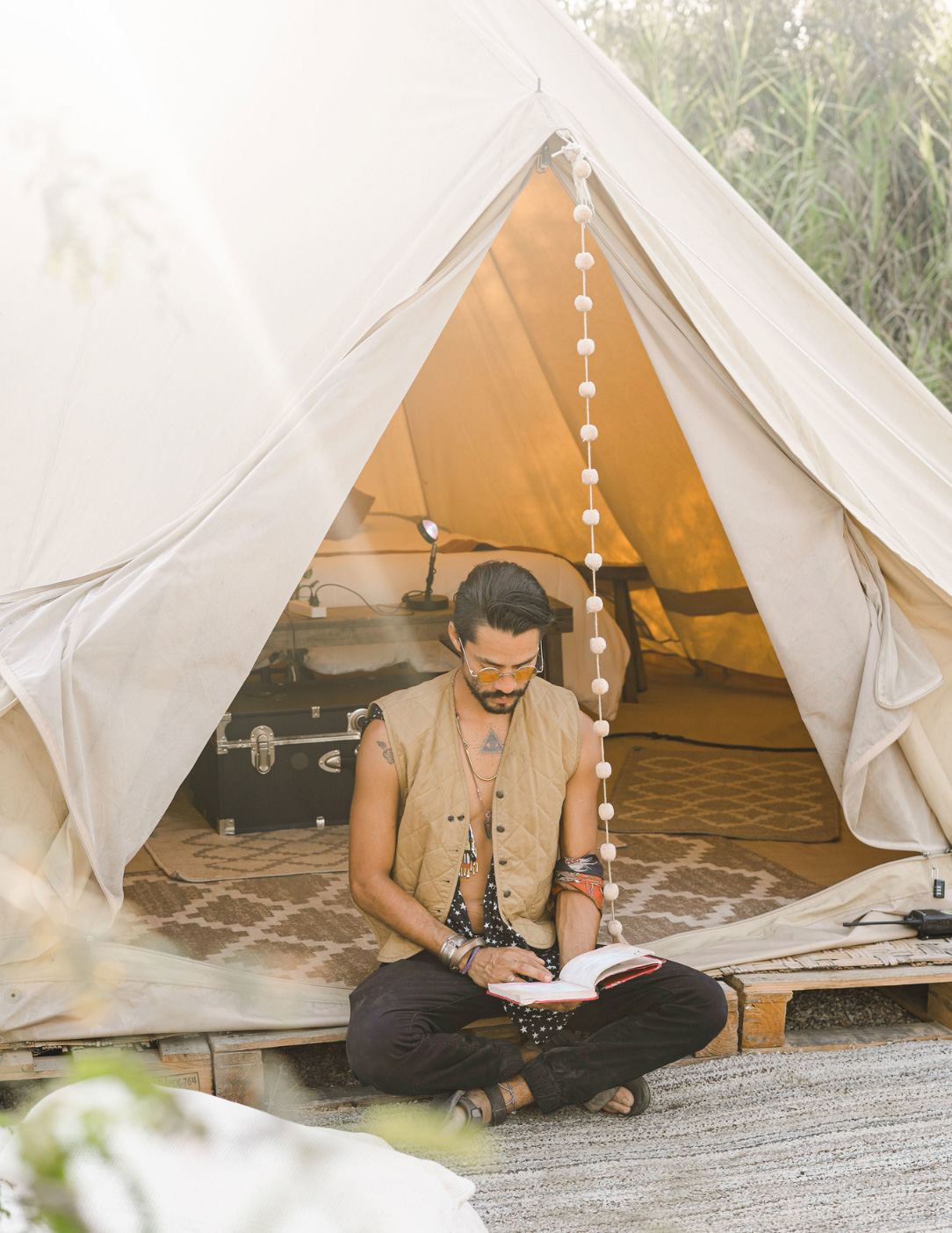 Man with sunglasses and tattoos sitting cross-legged reading a book in front of a beige tent.