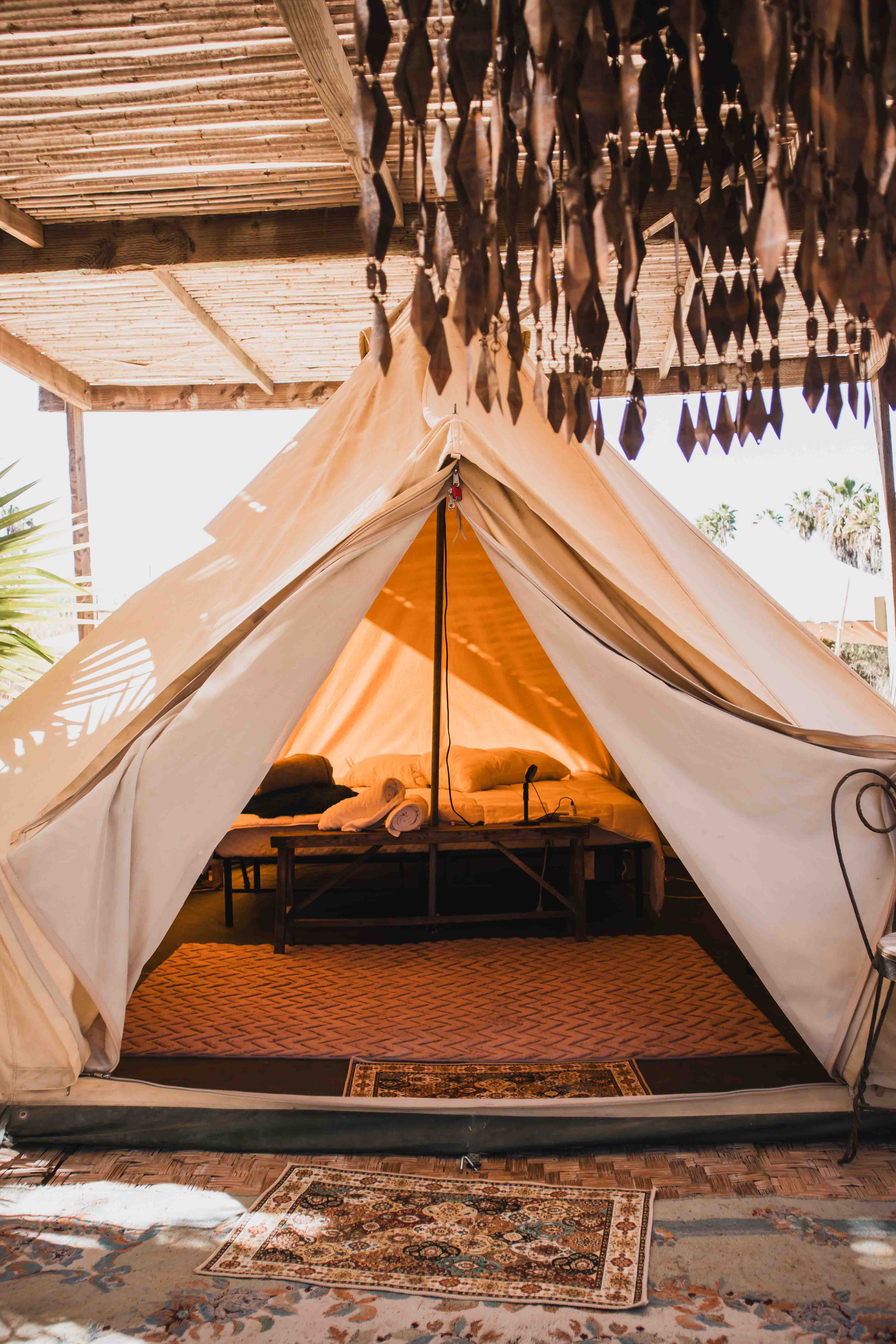 Interior view of a beige canvas tent with a bed, rolled towels, and a wooden bench under a bamboo roof.