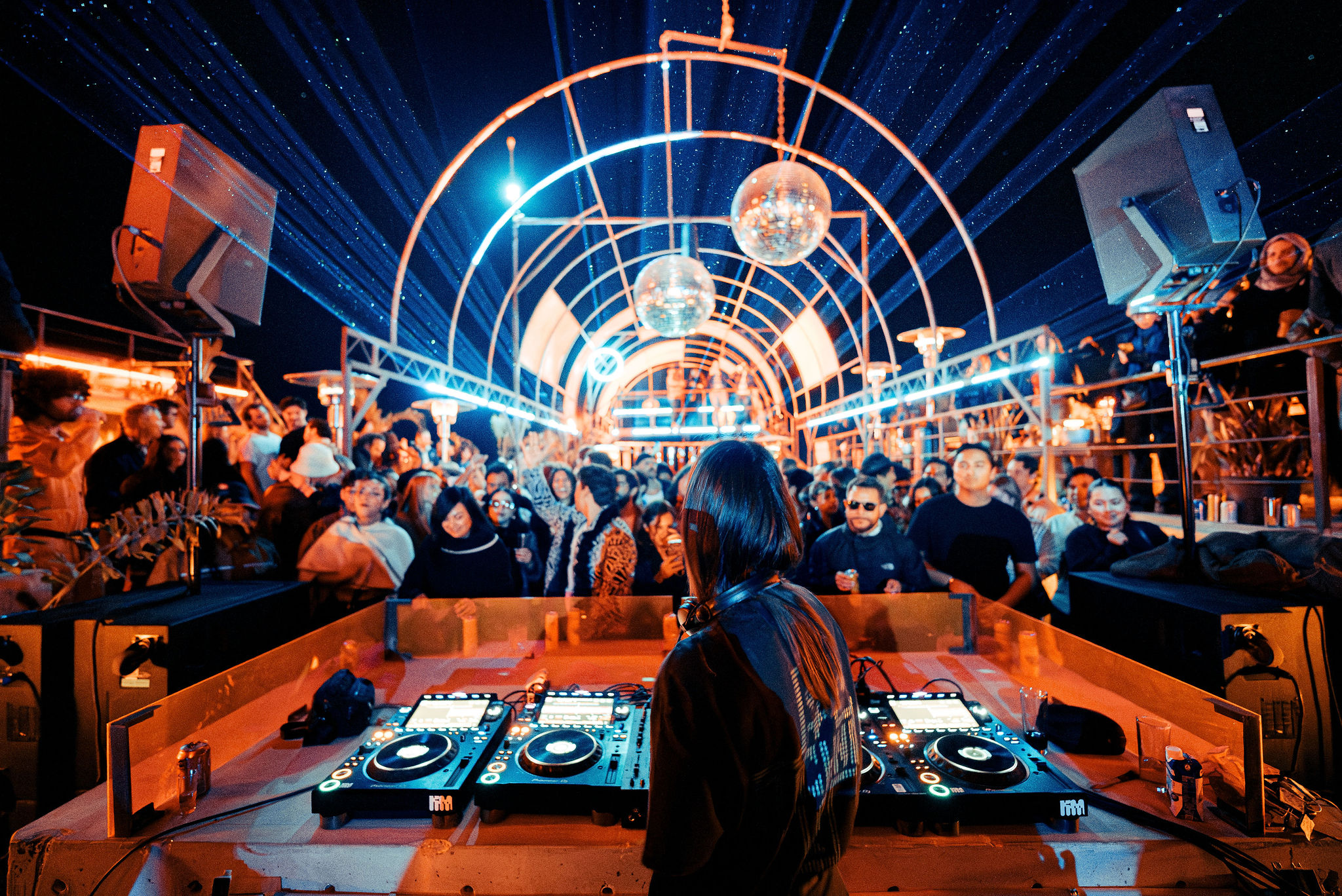 DJ performing at a vibrant outdoor night party with a crowd dancing under illuminated arches and disco balls.