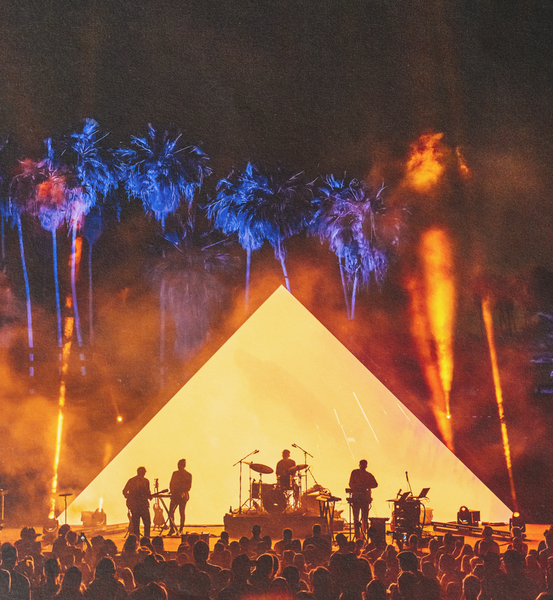 Silhouetted band performing on stage with a large illuminated triangular backdrop and blue-lit palm trees in the background at night.