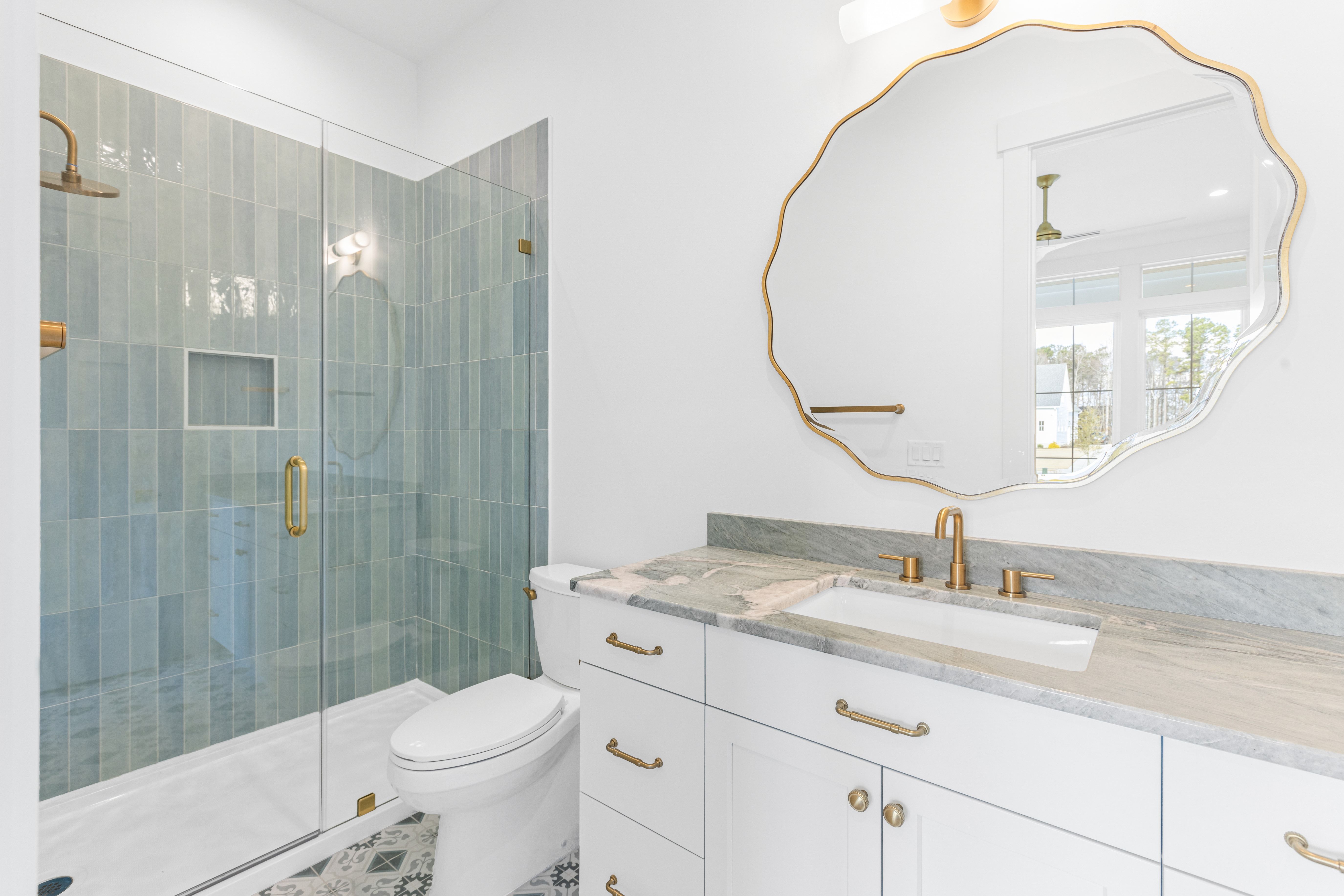 Bathroom with a wavy gold mirror, white vanity and green tile glass shower in the New Port custom home, Hampstead, NC