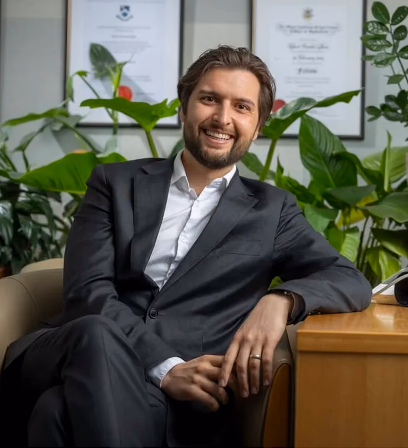 Man in suit smiles, sitting in chair with plants and certificates