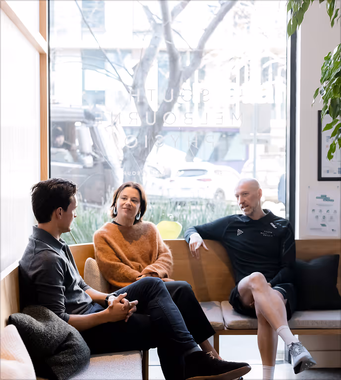 Three people sit on a bench in front of a large window