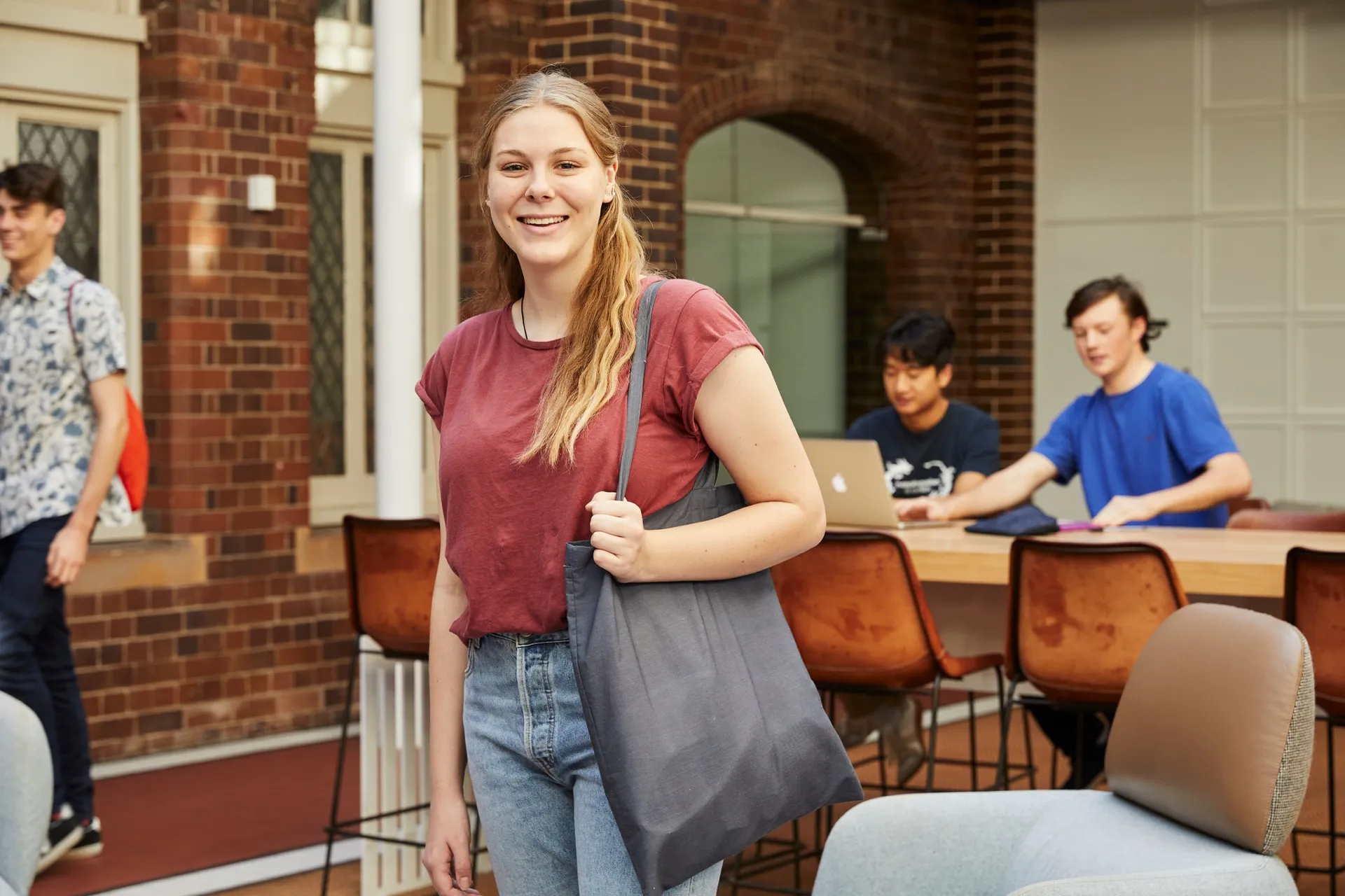 Smiling young woman with a long ponytail, wearing a red t-shirt and jeans, standing inside a brick building holding a gray tote bag, with two young men working on a laptop at a table in the background.