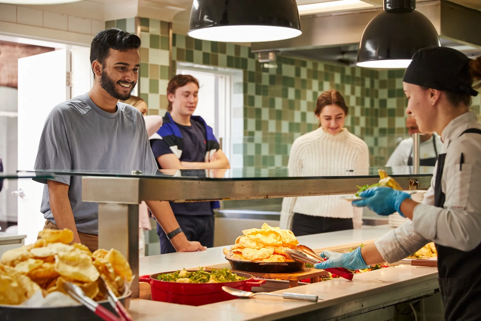 People waiting in line at a cafeteria counter as a server wearing gloves serves food.