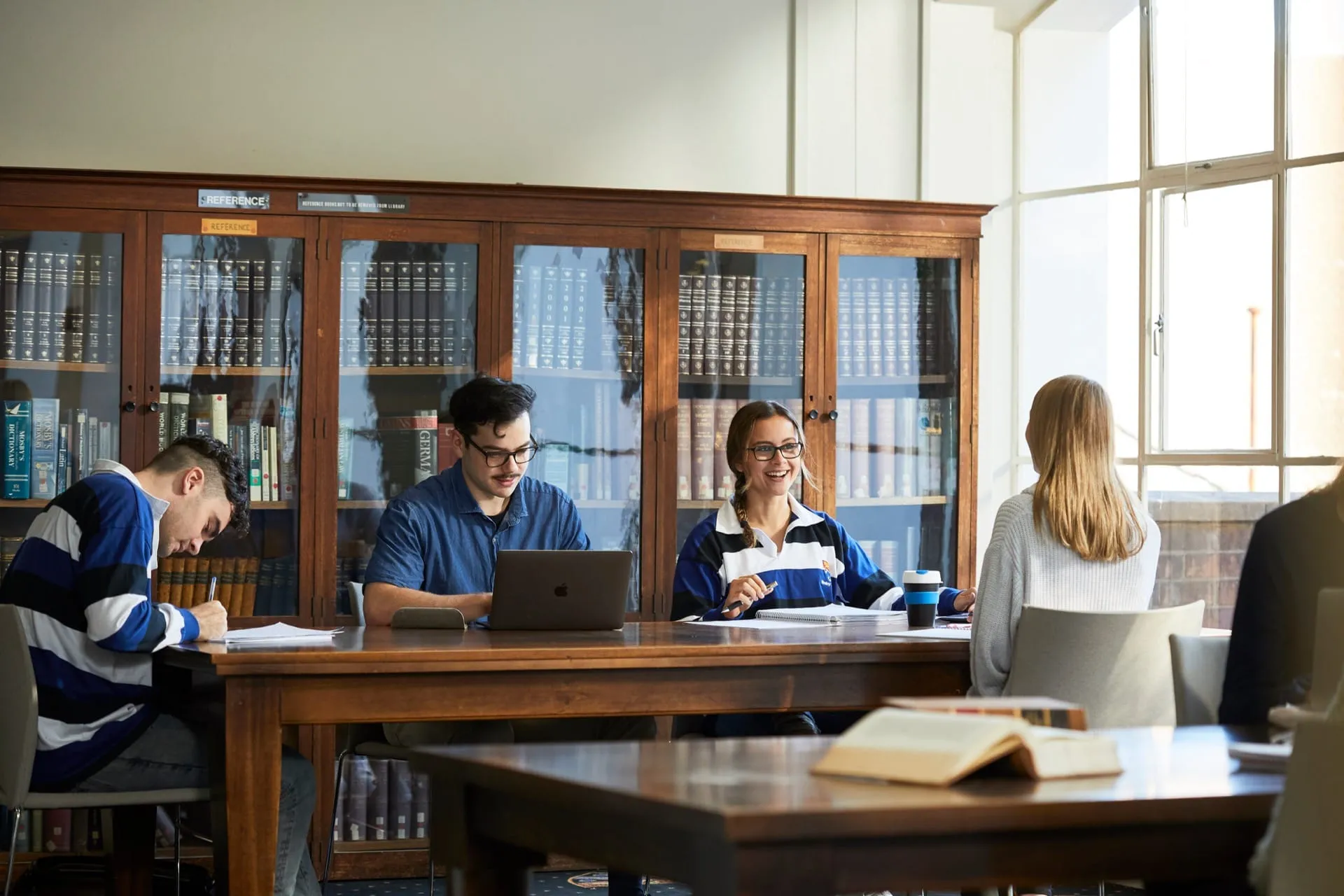 Group of four students studying together at a wooden table in a library with bookshelves and large windows in the background.