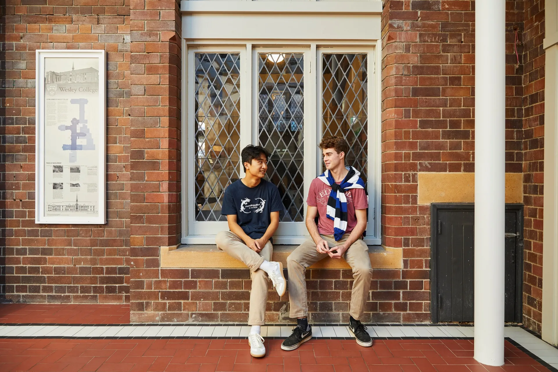 Two young men sitting on a brick ledge in front of a window with diamond-patterned glass, engaged in conversation.