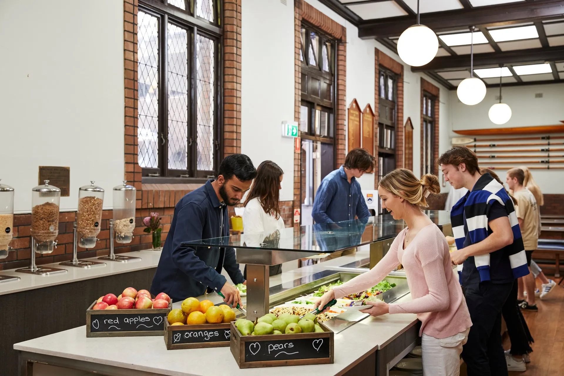 Several people selecting fresh fruit and salad from a buffet counter in a bright dining area with large windows.