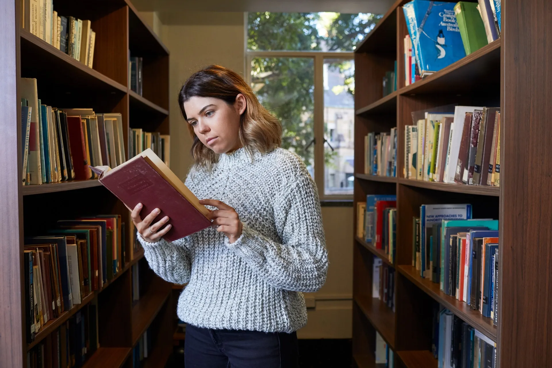 Young woman in a gray sweater reading a book between two bookshelves in a library.