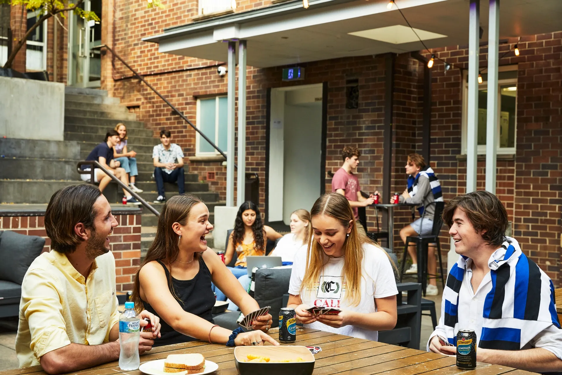 Group of young adults playing card games and socializing at an outdoor table with snacks and drinks.