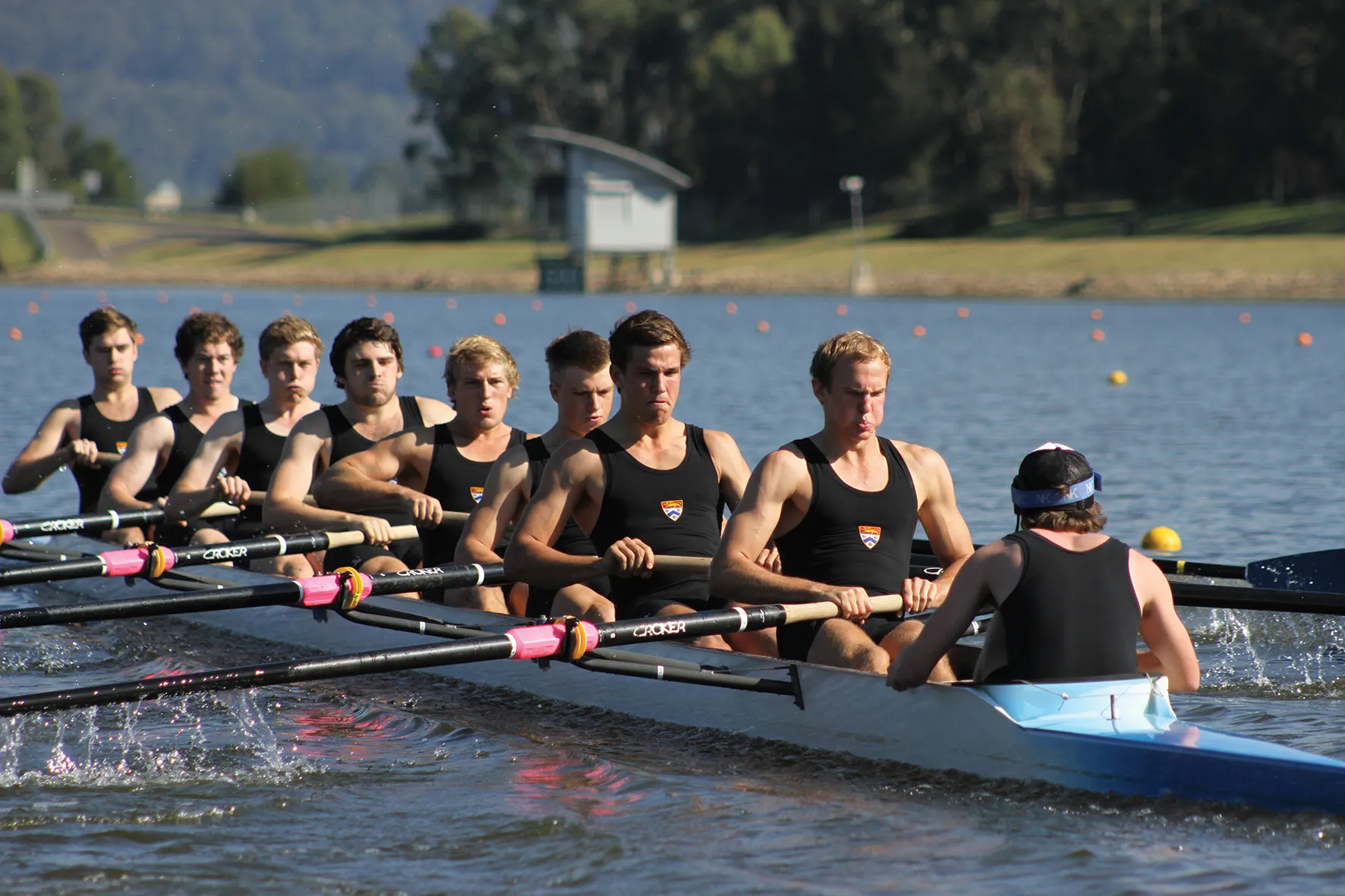 Rowing team of eight men rowing in sync on a calm lake with trees and a small building in the background.