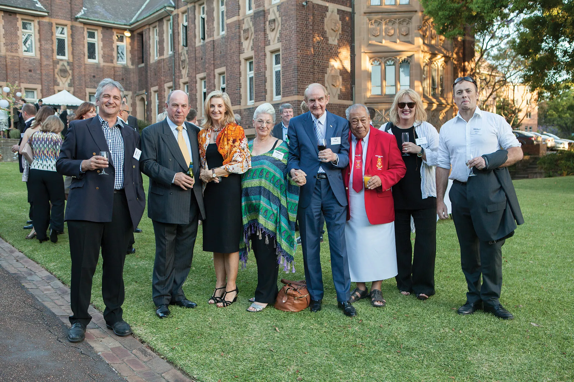 Group of eight well-dressed people standing on lawn outside historic brick building, holding drinks and smiling.