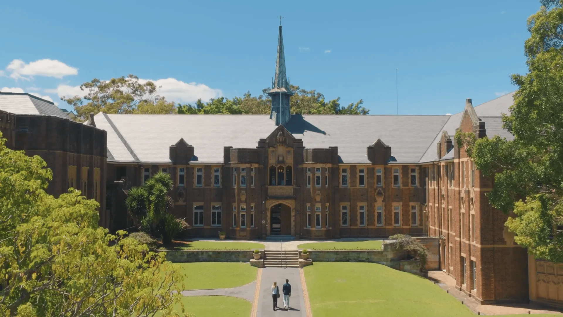 Two people walking on a path toward a large historic brick building with a central spire and surrounded by green lawns and trees.