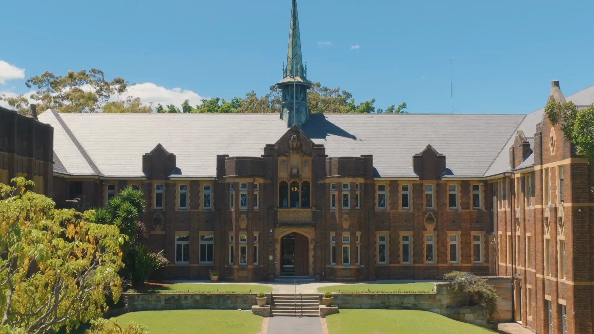 Historic brick college building with a central spire, surrounded by green lawns and trees under a clear blue sky.