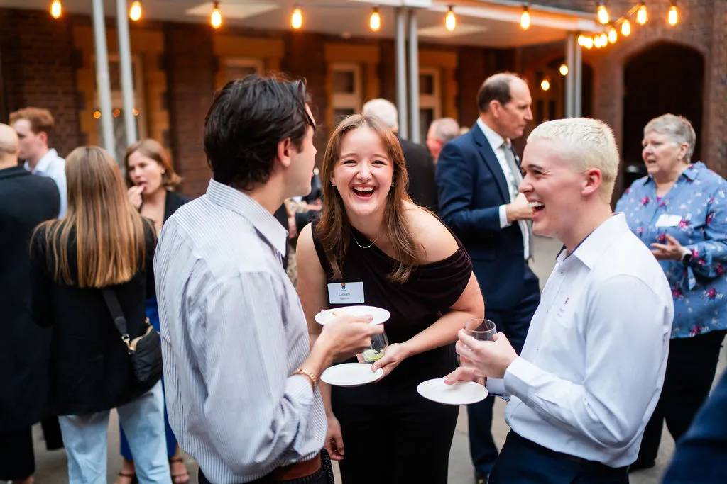 Three young adults smiling and laughing while holding plates and drinks at a social event with other guests in the background.