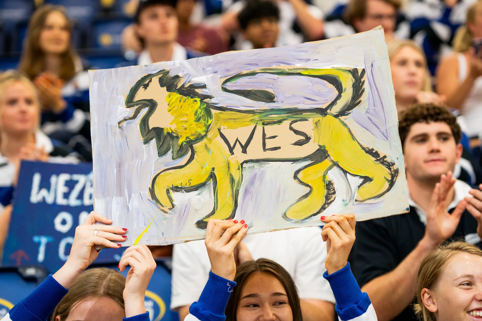 A person in a crowd holding a hand-painted sign of a yellow lion with the word WES written inside it.