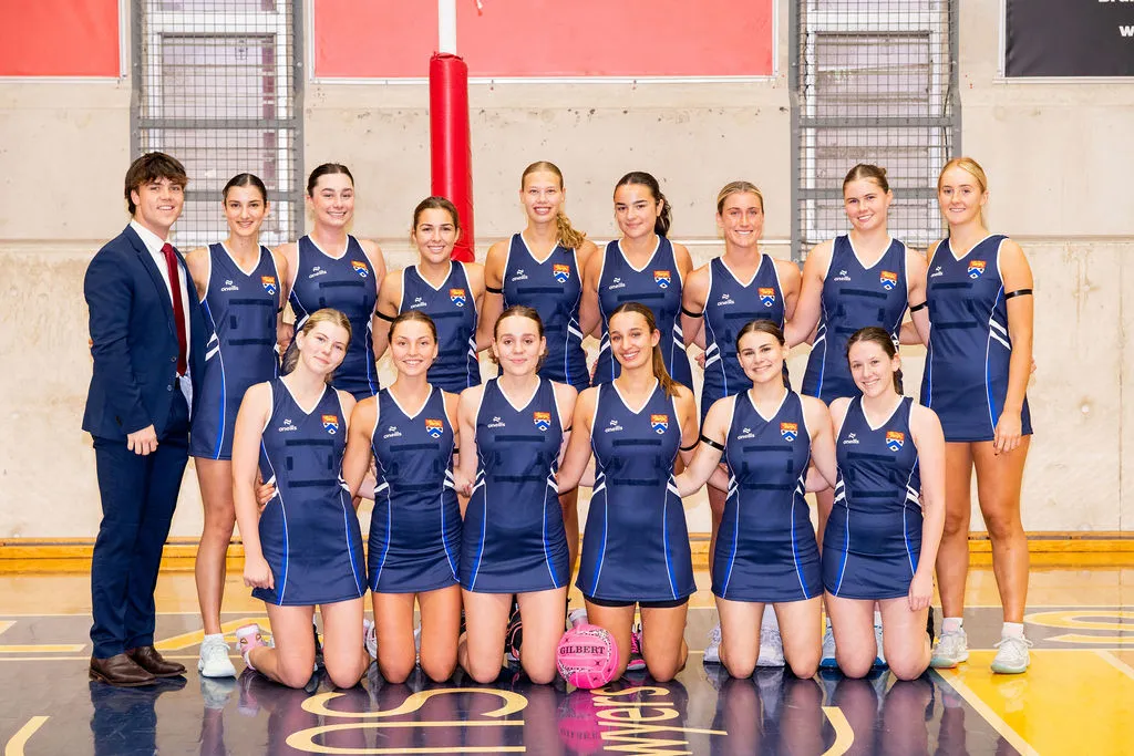 A women's netball team posing indoors in blue uniforms with a pink netball ball, accompanied by a man in a blue suit and red tie.