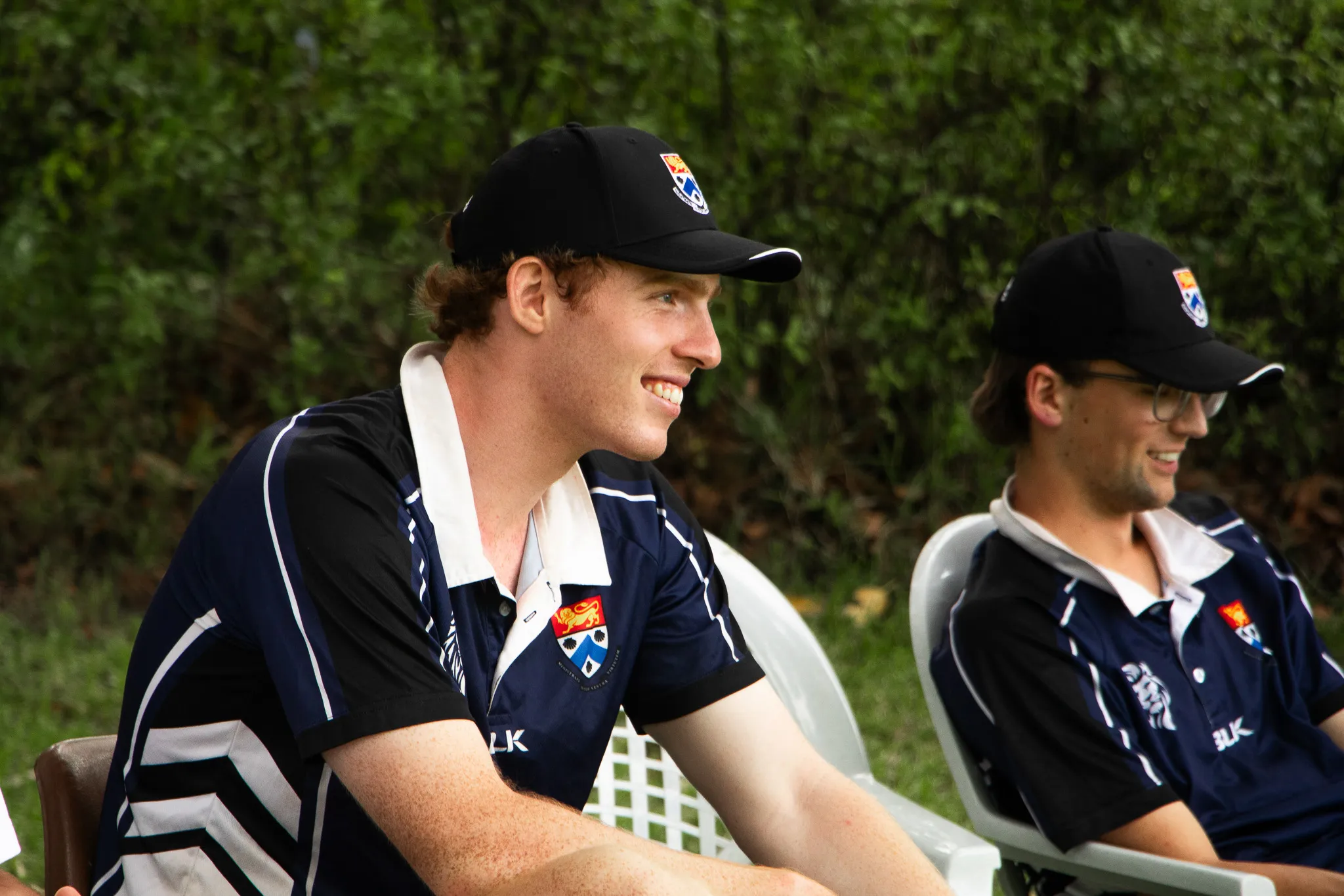 Two young men in matching navy and black sports uniforms and black caps sitting on white plastic chairs outdoors.