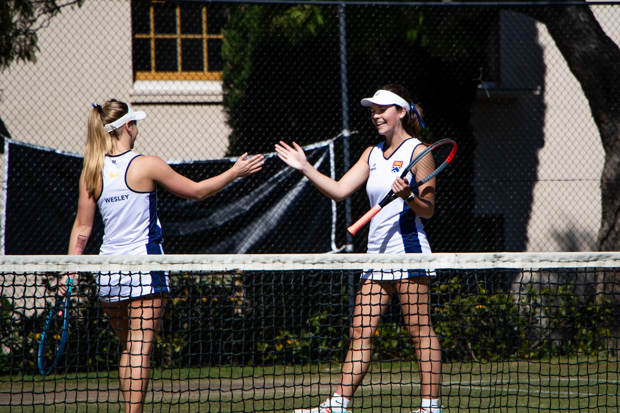 Two female tennis players in white uniforms smiling and giving a high five on a tennis court.