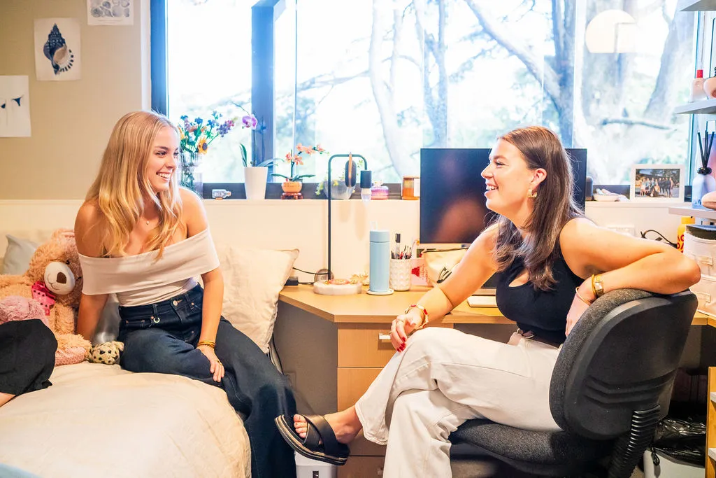 Two young women sitting and smiling in a bright, cozy dorm room with a teddy bear on the bed and a computer on the desk.