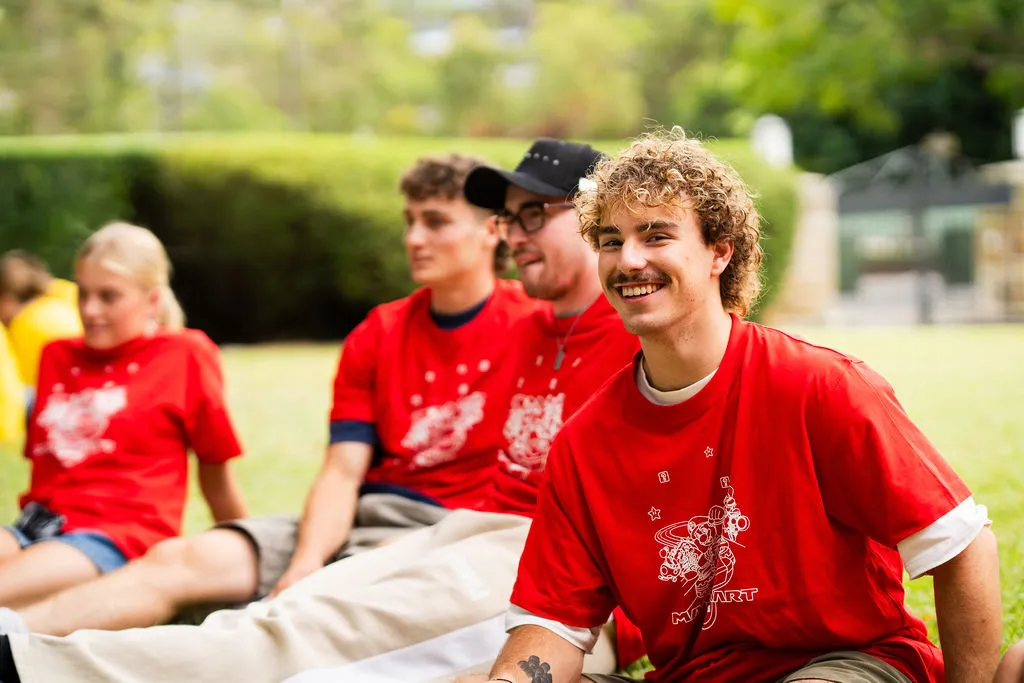 Four young people wearing matching red shirts sitting on grass outdoors, one smiling at the camera.