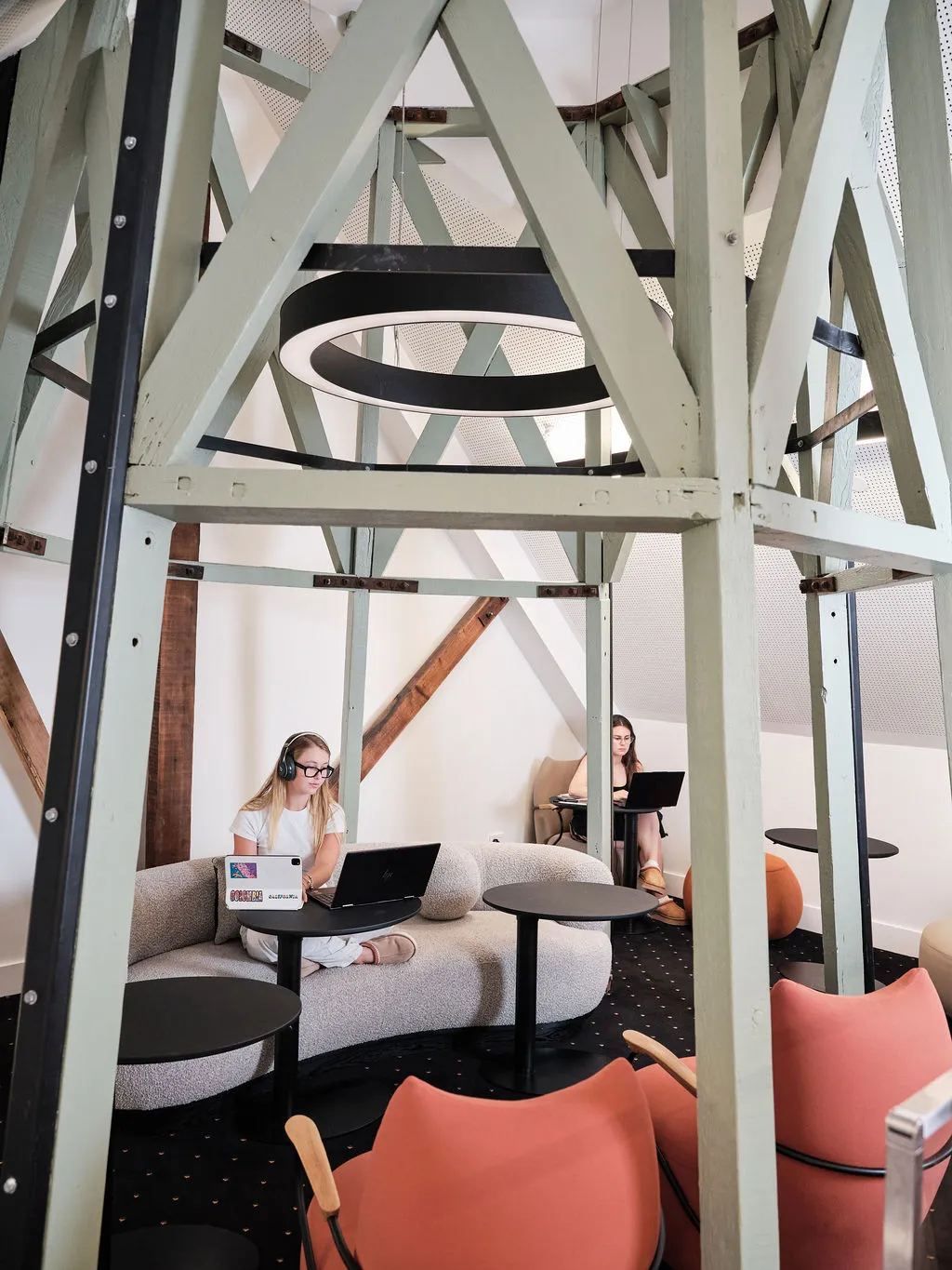 Two women working on laptops in a modern room with exposed beams and circular hanging light fixture.