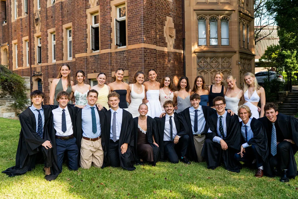 Group of young people dressed in semi-formal attire and graduation gowns posing on grass outside a historic brick and stone building.